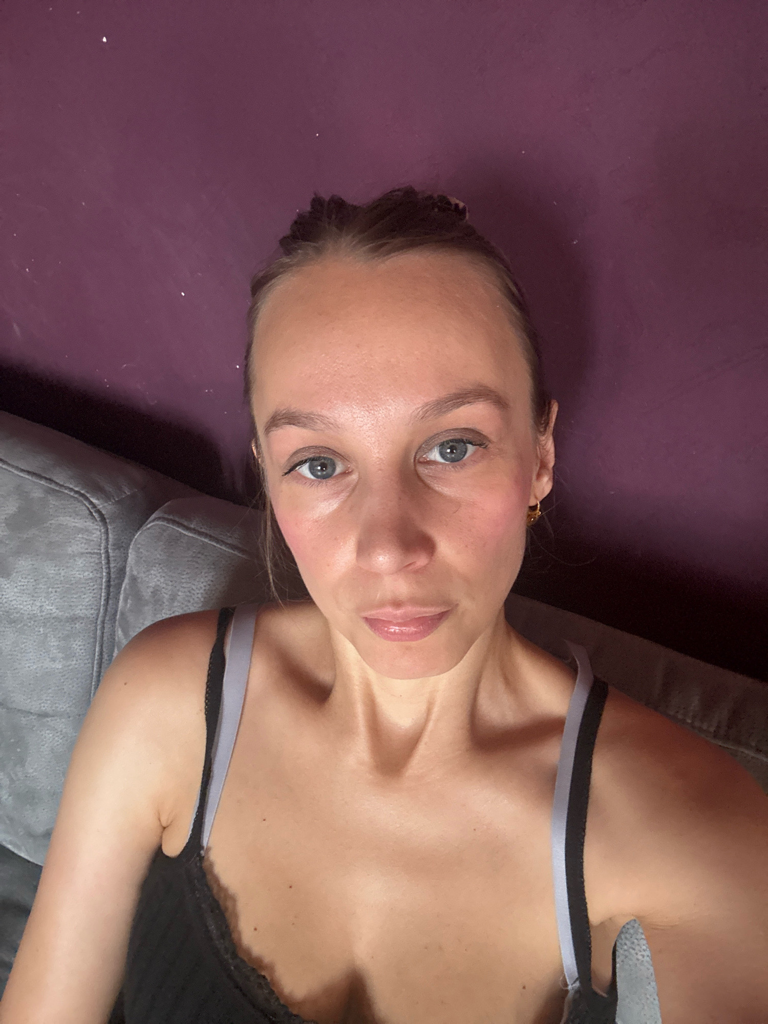 The image shows a close-up, overhead shot of a woman looking directly at the camera. She has light skin, light blue eyes, and her hair is styled in two braids. She is wearing a black tank top with grey straps. The background consists of a textured purple wall and a grey couch. The setting does not provide any specific clues to indicate the location in the city of Überherrn, Germany.