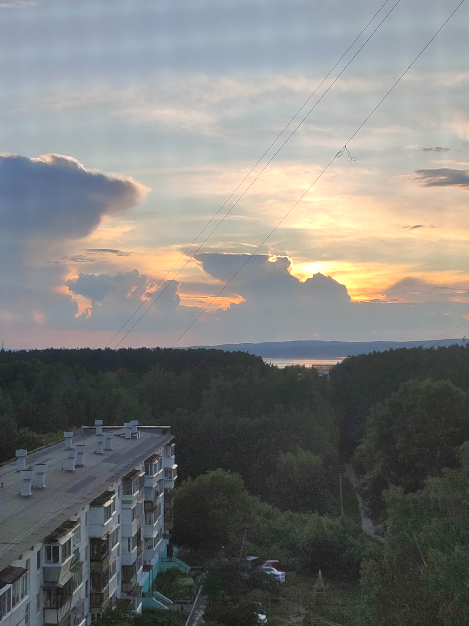The image captures a sunset scene over a body of water, with a dense forest in the foreground and a multi-story apartment building to the left. The sky is filled with dramatic clouds illuminated by the setting sun, casting warm hues of orange and yellow. Power lines stretch across the sky, with a small loop of material hanging from one of them. The apartment building, a typical Soviet-era structure, is flat-roofed and features numerous balconies and ventilation shafts. The forest appears lush and green, obscuring much of the landscape beyond the trees. A road is visible winding through the trees in the middle ground. A few cars are parked in a clearing within the trees. The overall impression is one of a peaceful, natural setting juxtaposed with urban development.