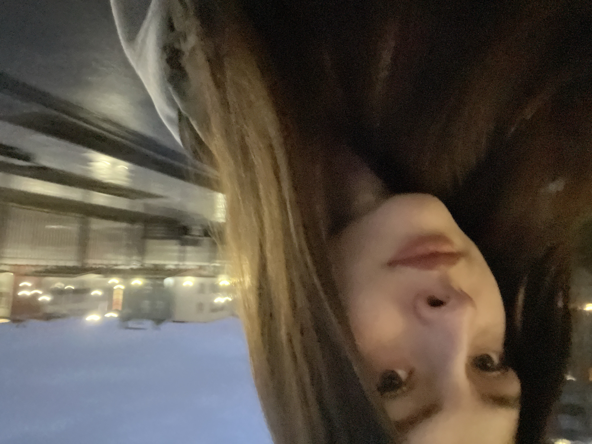 The image shows a person's face in close-up, with part of their hair visible. In the background, out of focus, is a snowy cityscape with lights. Based on the architectural style of the buildings and the presence of snow, the location is likely Saint Petersburg, Russia. Buildings and lights are reflected on a snowy ground. The face is tilted to the side.