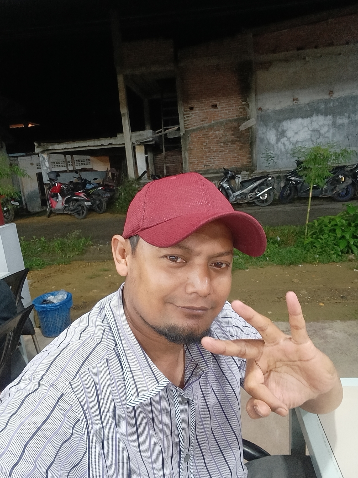 A man is photographed outdoors in the evening, seemingly in Lhokseumawe, Indonesia, evidenced by the architecture and environment. He wears a red baseball cap and a striped shirt. He is making a hand gesture with three fingers extended. Behind him are parked motorcycles, a partly constructed building, and greenery. A blue wastebasket is also visible.