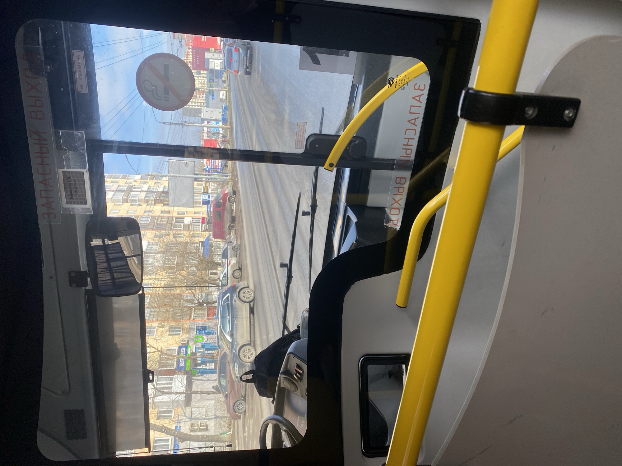 **Location:** Interior of a bus, likely in Izhevsk, Russia. The view through the windshield shows a city street with buildings, cars, and street signs that are consistent with Russian urban environments.
**Objects:**
* Bus interior elements: windshield, steering wheel, yellow grab bars, rear-view mirror, dashboard.
* Exterior view: buildings, cars, street, street signs, "No Smoking" sign on the windshield.
**Texts:**
* A “No Smoking” sign on the windshield.
* The text “ЗАПАСНЫЙ ВЫХОД” (“Emergency Exit”) is seen on the left side.
* The text “ЗАПАСНЫЙ ВЫХОД” (“Emergency Exit”) is also visible on the right side (mirrored), and other text is also present.
**Environment:** Urban setting with moderate lighting conditions. It appears to be daytime.