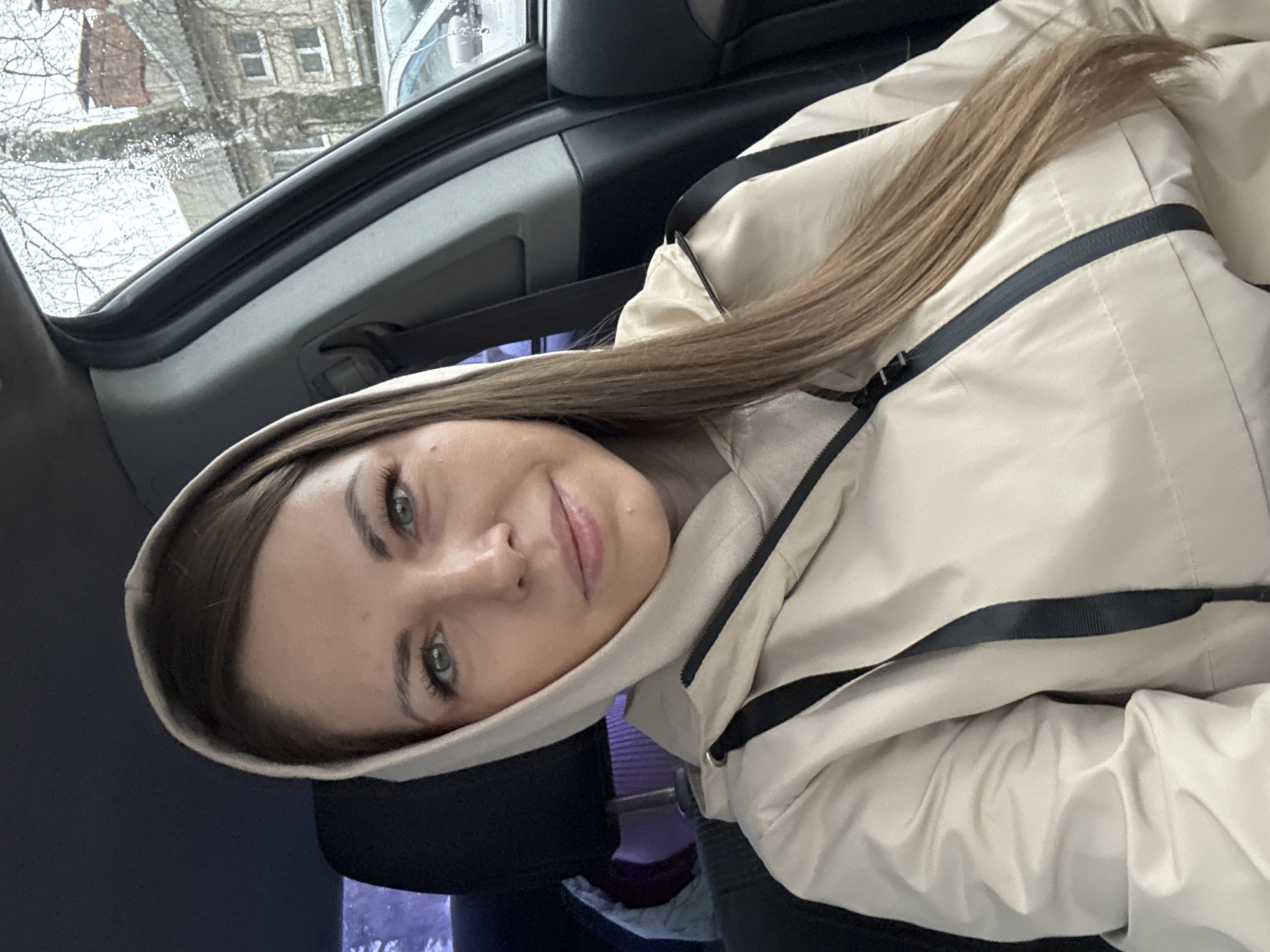 A young woman is sitting in a car, wearing a beige hooded jacket. She has long brown hair and green eyes. She's looking at the camera with a slight smile. Outside the car window, a cityscape is visible, possibly in Razdolye, Russia. The buildings appear to be covered in snow or ice.
