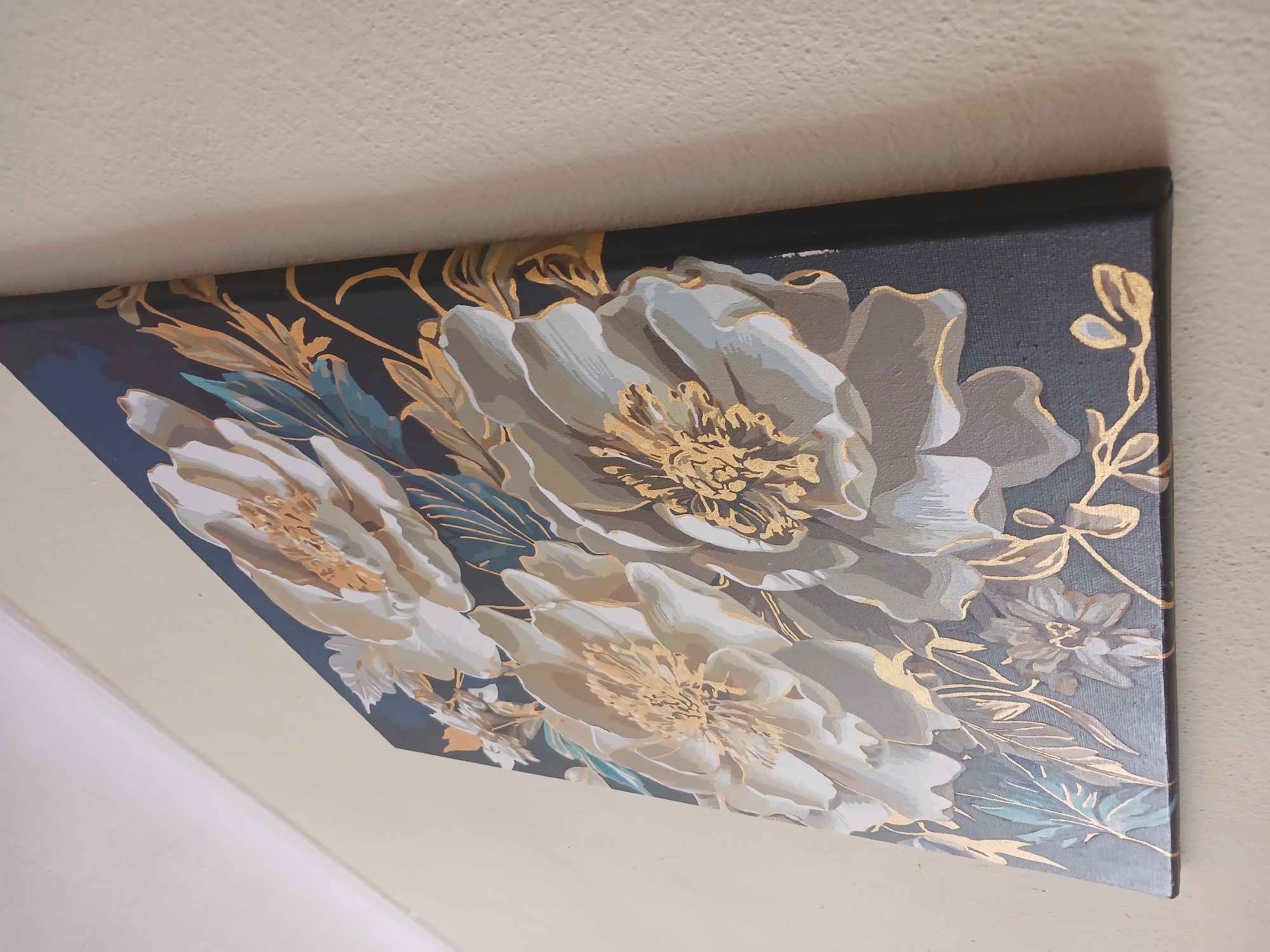 A painting of three large, light-colored flowers with gold accents is hanging on a cream-colored wall. The painting is angled, with the top left corner higher than the bottom right. The flowers have detailed gold centers and highlights on their petals. Surrounding the flowers are gold leaves and some blue-green foliage. The background of the painting is a dark navy blue. The wall appears to be textured. The scene is indoors, suggesting it is in a room within a building. There are no people visible in the image.