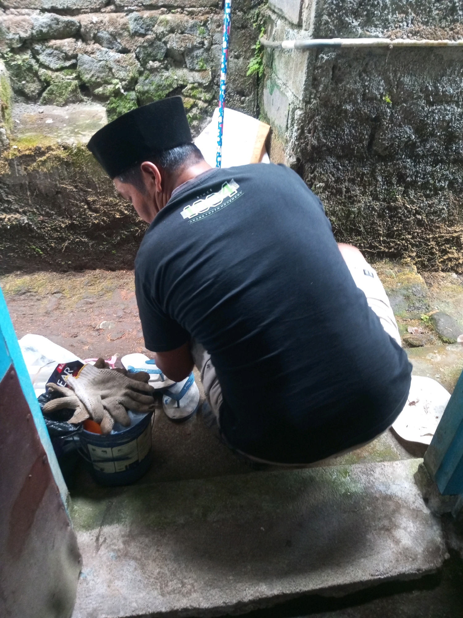 A man wearing a black peci and a black t-shirt is squatting on a concrete step. He is facing away from the camera, and his head is bowed. In front of him is a blue bucket with a black bag inside. The bag contains a pair of brown work gloves and an orange object. There are also a pair of blue and white flip-flops to the right of the bucket. The background is a stone wall with moss growing on it. The scene appears to be outdoors, possibly in a residential area in Andongsili, Indonesia, given the style of the stonework and the presence of tropical vegetation.