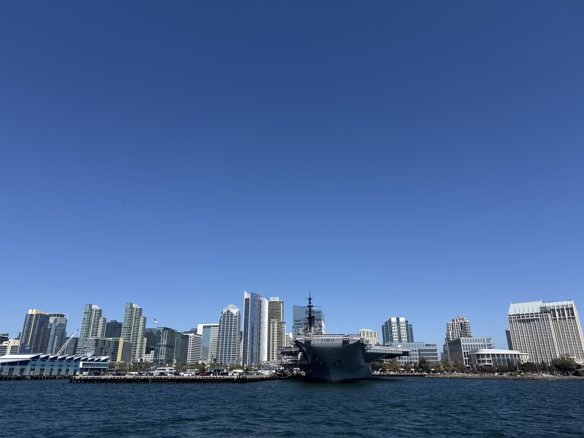 This is a wide-angle, eye-level shot of the San Diego, California skyline taken from the water on a clear, sunny day. In the foreground, the dark blue, choppy water of San Diego Bay stretches across the bottom of the frame. Dominating the midground is the massive gray hull of a docked aircraft carrier, identified by the name "MIDWAY" on its stern. The carrier museum appears to be a significant attraction, with its bridge structure and deck clearly visible.
Behind the aircraft carrier, the San Diego skyline rises, featuring a variety of modern skyscrapers in shades of glass, white, and gray. Some buildings have distinct architectural designs, including curved facades and stepped tops. Visible on the waterfront behind the carrier and to the left are piers and buildings associated with the Embarcadero, including a long, low building with a blue roof and a prominent crane. Cars and buses are parked and driving along the waterfront. The sky above is a clear, vibrant blue with no clouds. The overall scene depicts a bustling waterfront with a prominent historical landmark against the backdrop of a modern urban landscape.