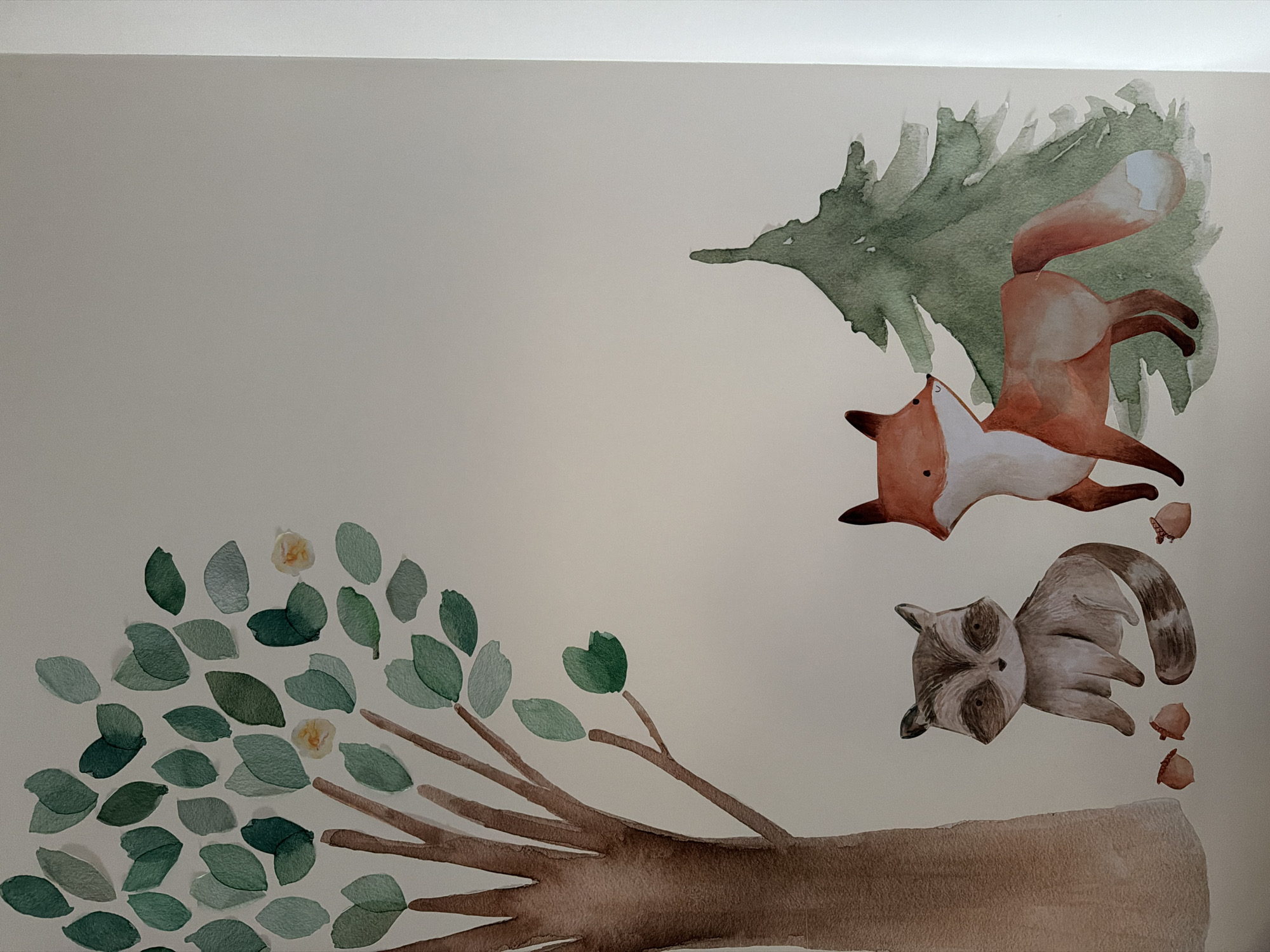 The image shows a watercolor painting on a light beige wall. The painting depicts a whimsical forest scene with a fox leaping near a green fir tree, a raccoon, acorns, and a tree branch with leaves and small yellow flowers. The setting does not appear to be located in Moscow, Russia.