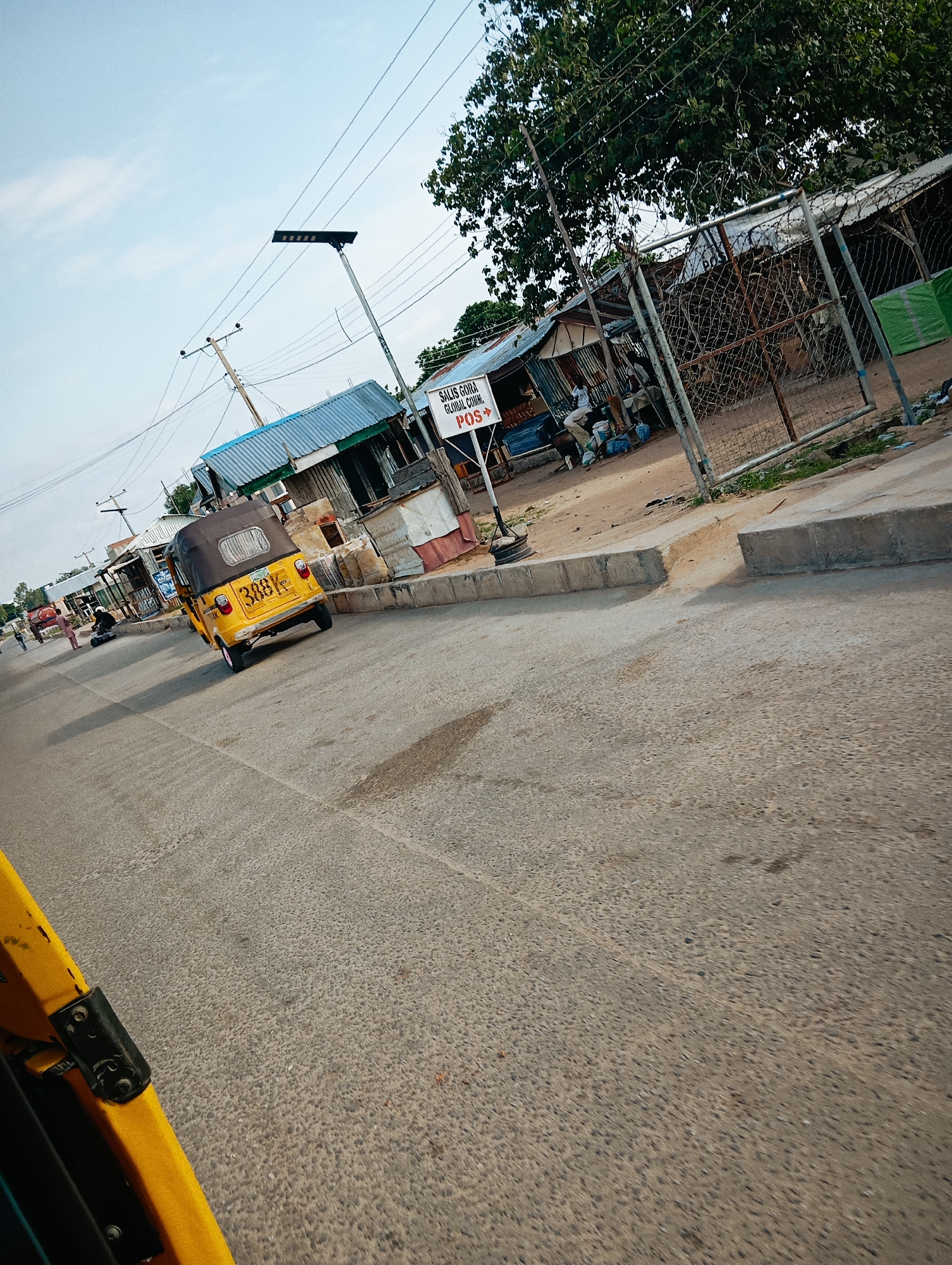 The image captures a street scene in Jalingo, Nigeria, likely a commercial area given the presence of several small shops. In the foreground, a yellow auto-rickshaw, also known as an "okada" or "keke napep," is turning on the road. Its license plate, though partially obscured, reads "388K". The road surface is asphalt, showing signs of wear and tear.
To the right of the road, a sidewalk runs alongside a row of businesses. Some of these structures have corrugated metal roofs, characteristic of many informal markets and shops in Nigeria. A prominent white sign with red lettering is visible, indicating "SALIS GHANA GLOBAL COMM. POS." This suggests a communication or business services provider. Further down the street, a fence made of chain-link and barbed wire encloses a property, behind which more corrugated structures and what appear to be tents or tarpaulins are visible.
In the background, utility poles with a network of wires stretch across the sky, typical of urban infrastructure. A solar-powered street lamp is mounted on one of the poles. A few individuals are present near the shops, some appearing to be customers or vendors. The overall atmosphere suggests a busy, everyday urban environment. The sky is a pale blue with some wispy clouds, indicating daytime. The image has a slightly tilted perspective, possibly taken from a moving vehicle.