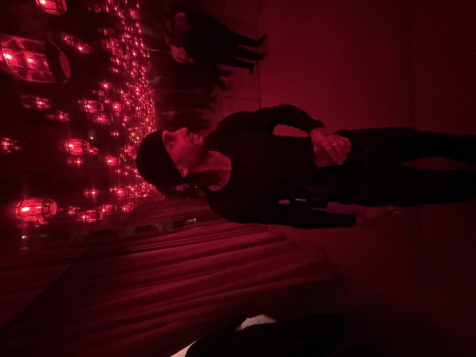 The image captures an indoor scene dominated by deep red illumination, consistent with the distinctive "red room" interior of the Gucci Osteria da Massimo Bottura in Milan, Italy. A male individual, estimated to be an adult, stands slightly right of the frame's center, facing towards the right with his gaze directed off-camera. He is dressed in a black beanie, a long-sleeved black shirt, and dark trousers. He holds a transparent glass containing a reddish liquid in his left hand, on which a ring is visible. His right hand is not fully visible but appears to be at his side or hip. To the immediate left, a wall covered in red curtains or fabric is present. The ceiling above is adorned with numerous glowing red lantern-style light fixtures, which are the primary source of the pervasive red light. In the blurred background on the right, several indistinct human figures are visible, also enveloped in the strong red light. The floor is uniformly red and lacks specific discernible features.
