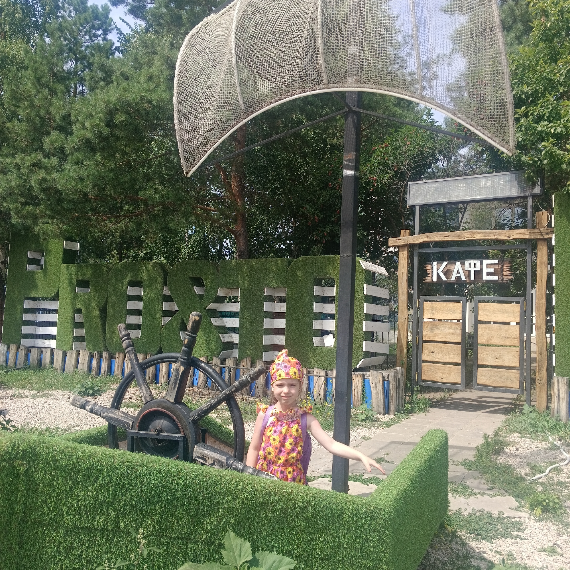 A young girl in a colorful floral dress and headscarf stands to the right of a ship's wheel, which is encased in a low, green, artificial grass border. She is pointing with her right hand towards the viewer. Behind her, large green letters spelling "ПАРК" (PARK in Russian) covered in artificial grass are visible, backed by white wooden planks. To the far right is an entrance with a sign that reads "KATE" in English, with wooden double doors below. The scene appears to be outdoors, with trees and foliage in the background, suggesting a park or recreational area in Novokuybyshevsk, Russia.