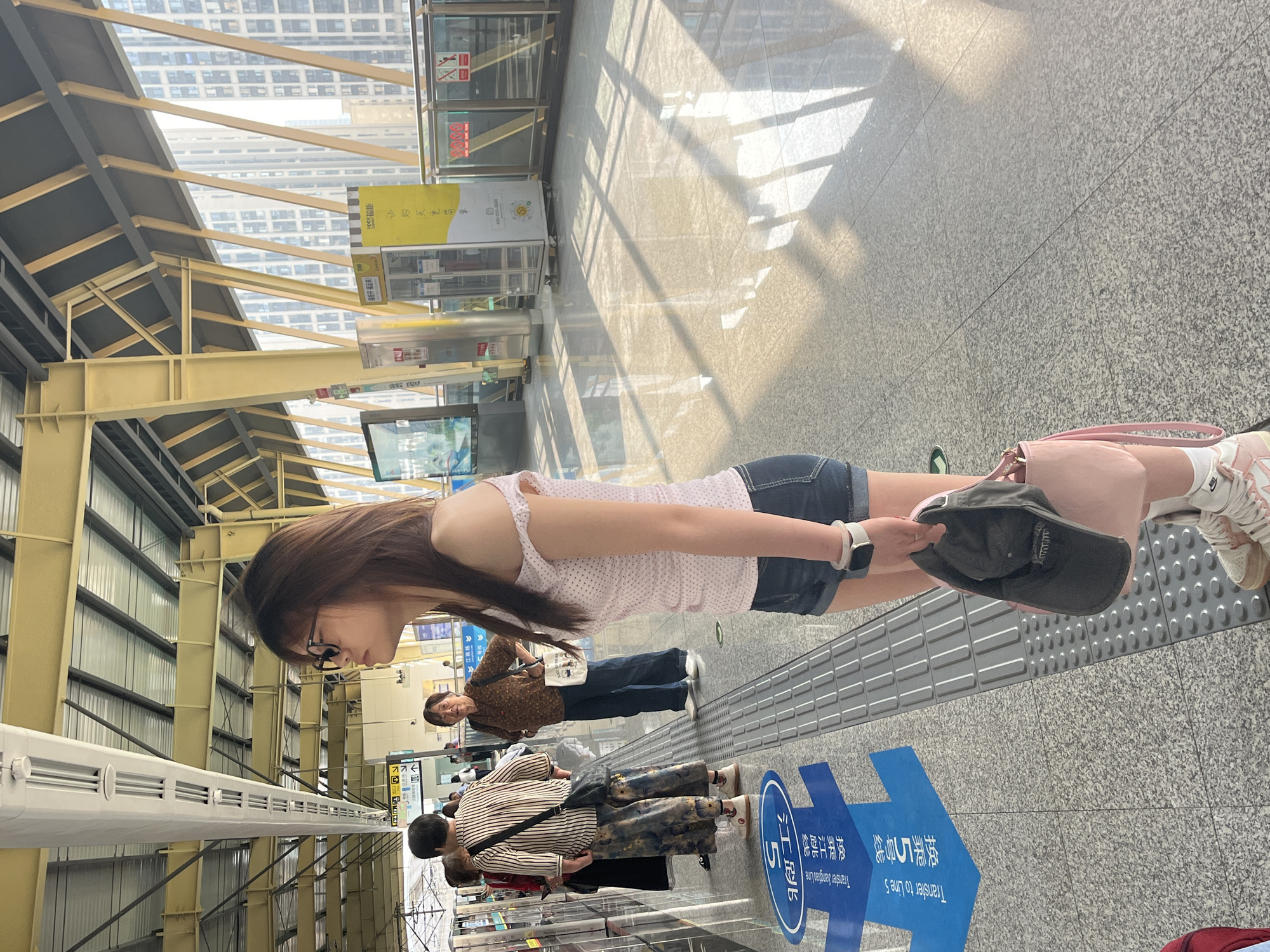 A young woman with shoulder-length brown hair and glasses stands on a brightly lit train platform. She is wearing a pink off-the-shoulder top with white polka dots and denim shorts. She is holding a grey cap and a pink handbag. She has a watch on her left wrist. The platform is made of speckled grey tile and has a tactile warning strip along the edge. A blue directional sign indicating "Transfer to Line 5" is visible on the floor in front of her. In the background, other passengers are waiting, and a modern train station structure with yellow beams and a translucent roof is visible. The scene is likely in the city of Tiaodeng, China, given the signage in Chinese characters.