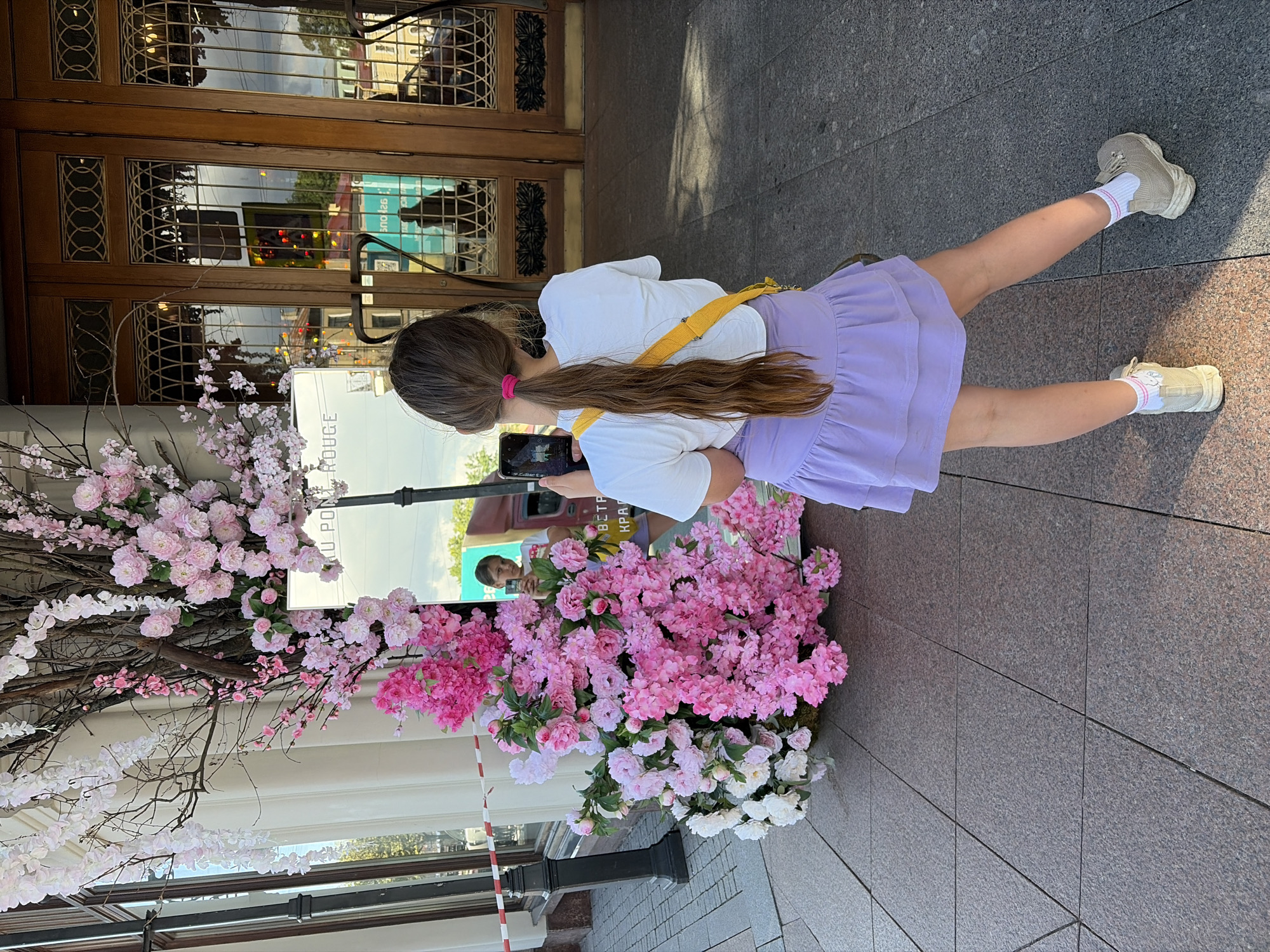 A young girl with long brown hair tied back in a ponytail is standing outside, taking a photo with her phone. She is wearing a white t-shirt, a lavender skirt, white socks with pink stripes, and beige sneakers. A yellow strap is visible across her chest, likely from a bag. The background features a building with large wooden doors and windows, reflecting the surroundings. In front of the building, there is an elaborate floral display with numerous pink and white cherry blossoms and greenery. The ground is paved with grey granite tiles. The scene suggests a sunny day, likely in the spring or summer, given the blossoms. The girl appears to be casually taking a photo of the flowers or her reflection. Text visible on a sign reads "ROUGE" and "VPON". The location is Saint Petersburg, Russia.