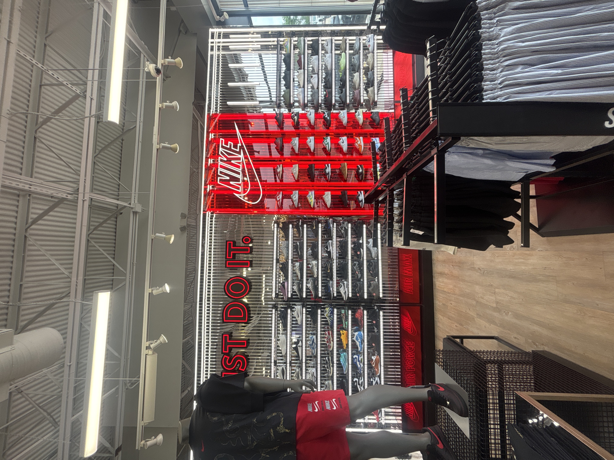 The image displays the brightly lit interior of a modern retail store, specifically a prominent Nike section. A large, red neon sign is centrally featured, vertically spelling "NIKE" and "JUST DO IT." This sign is integrated into a multi-tiered display system showcasing numerous pairs of athletic shoes, primarily sneakers, on individual shelves. Below the main neon signs, additional red text labels on the display panels indicate shoe categories such as "AIR FORCE" and "AIR MAX."
In the foreground, a mannequin is visible, dressed in a black short-sleeved top, red athletic shorts adorned with white Nike logos, and black and red Nike sneakers. To the right of the central display, black metal shelving units hold neatly folded clothing, including stacks of blue and white striped shirts and dark solid-colored apparel. A black sign with a partially visible letter "S" is present on these shelves.
The store's architecture includes an exposed ceiling with corrugated metal panels, white structural beams, and a mix of long fluorescent strip lights and smaller directional spotlights, contributing to the bright and modern aesthetic. The flooring in the visible areas consists of light-toned wood laminate. The scene depicts an organized and inviting retail space for sportswear, consistent with a store found in Arlington, United States of America.