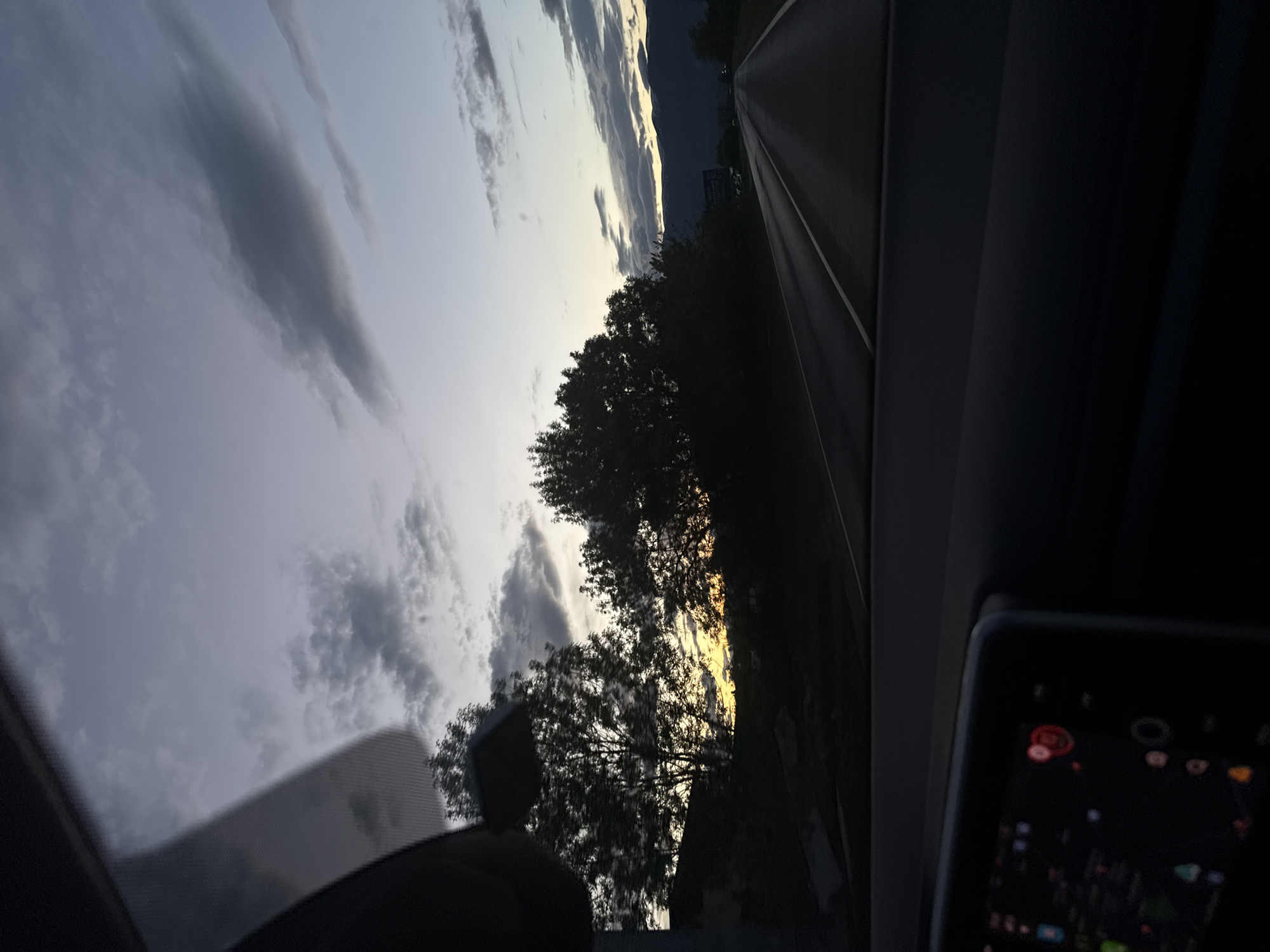 The image shows a view from inside a car, likely in motion, at dusk or dawn. The sky is filled with gray and white clouds, with a glimpse of yellow light behind trees suggesting sunrise or sunset. To the right, a road stretches out ahead, framed by the car's interior. A digital display screen is visible in the lower right corner. The location is likely a rural or suburban area given the road and trees.