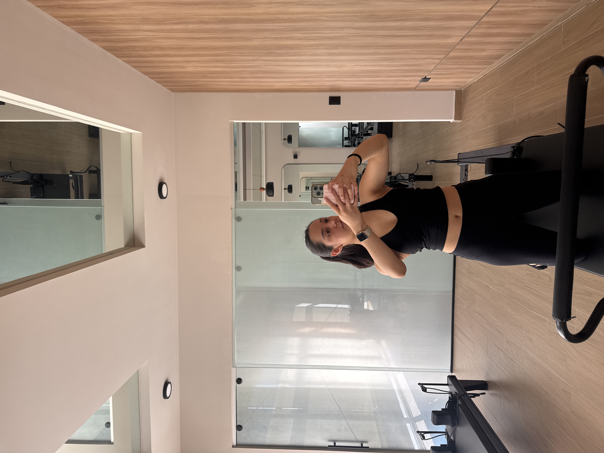 A young woman is captured taking a mirror selfie in what appears to be a modern Pilates or fitness studio. She has dark hair and is dressed in a black sports bra and black leggings, with a silver smartwatch on her left wrist. She holds a smartphone, encased in a light pink checkered cover, to her face, positioned to photograph her reflection.
The interior of the studio features clean, contemporary design elements. The walls are a light beige or off-white, complemented by a light wood-patterned ceiling and a matching tiled floor. The room is brightly lit, likely by recessed ceiling fixtures, and a circular black wall sconce is visible on the left wall. Large glass panels and mirrors line the back and side walls, creating reflections of the studio's interior. These reflections prominently display several black Pilates reformer machines, indicating the facility's purpose. One such reformer is partially visible in the foreground on the right. A notable architectural detail is a section of wall with horizontally striped frosted glass, adding texture and privacy to the reflections. The activity suggests a pause during or after a fitness session. There is no visible text within the image. The location context provided is San Bernardino Tlaxcalancingo, Mexico.