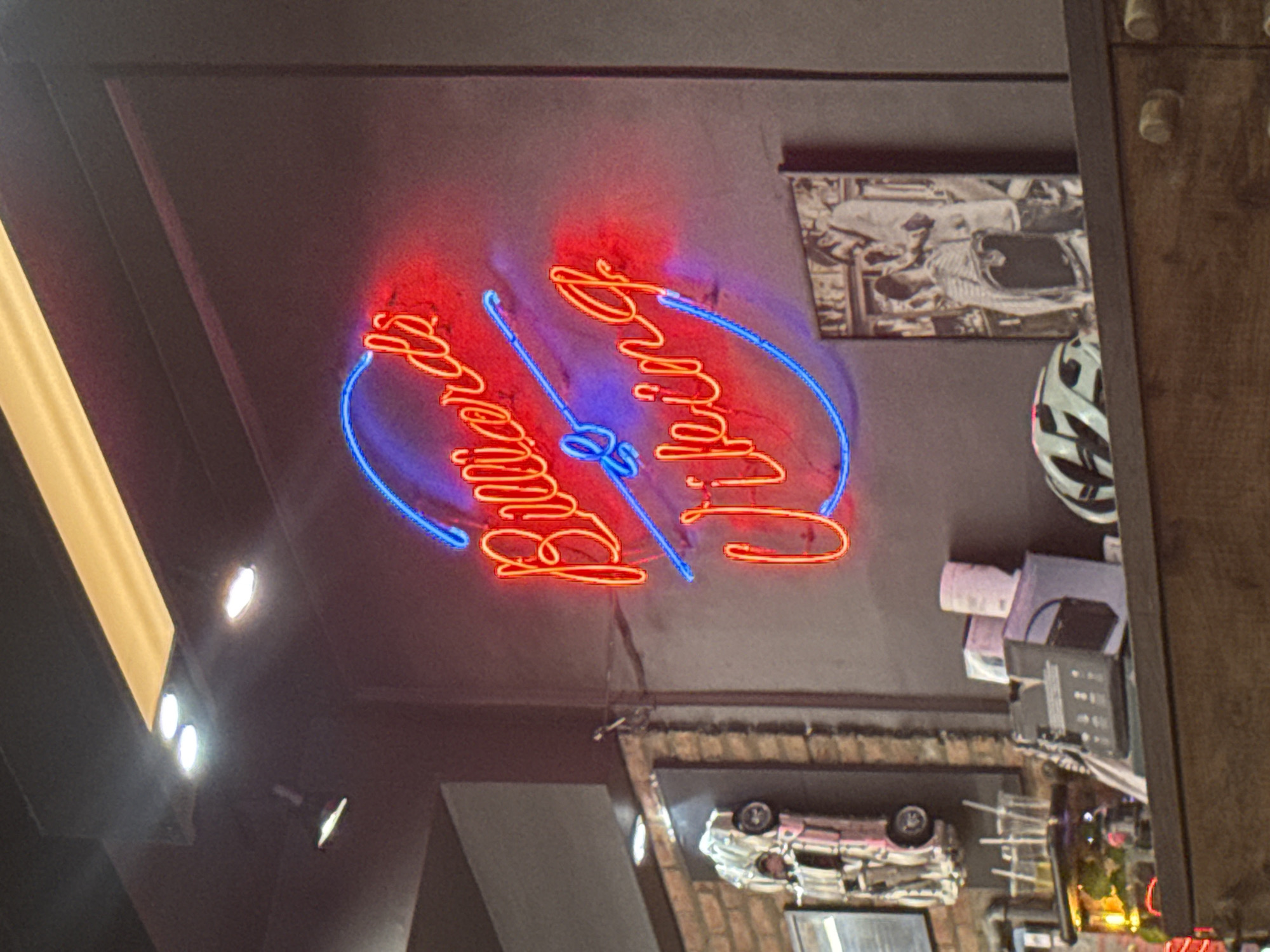 An interior view looking up at the ceiling. A neon sign that says "Burger Bros" is centered. The neon sign has a red glow for the letters and a blue glow for the design. Adjacent to the neon sign are decorative items, including a framed picture and a model car on display. Part of the ceiling is a dark gray. The image was captured at a Burger Bros restaurant in Đà Nẵng, Vietnam.