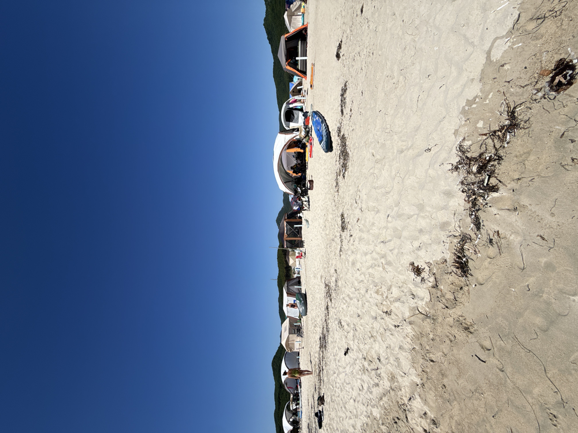 A wide, low-angle shot captures a busy beach scene with numerous tents and people under a clear blue sky. The foreground is dominated by sand with scattered seaweed. In the mid-ground, several tents of various sizes and colors are set up, creating a temporary campsite. Some tents are open, revealing people inside or sitting nearby. Inflatable watercraft, including a paddleboard and a small inflatable boat, are visible on the sand. A woman in a green swimsuit walks across the sand, and another woman in a black swimsuit is seated near an inflatable boat. In the background, a line of green, tree-covered hills forms the horizon. The overall impression is of a recreational gathering on a sunny day at the beach, potentially in the vicinity of Nakhodka, Russia, given the geographical context.