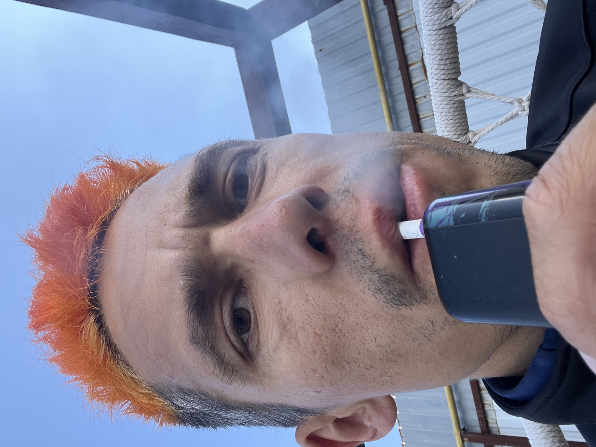 A low-angle selfie, rotated approximately 90 degrees counter-clockwise, depicts a man vaping. The man has distinctive bright orange hair, dark eyebrows, and stubble on his face. He looks directly at the camera with a neutral to slightly focused expression, his eyebrows subtly furrowed. He holds a black and purple vaping device to his mouth, with a white tip inserted, and a plume of white vapor or smoke is visible around his lips and chin. He is wearing a dark-colored jacket, possibly black or dark blue, over a blue layer.
The background on the left features a clear blue sky. To the right, various structures are visible, including light grey corrugated metal siding, dark wooden beams, and a section of white, tightly woven rope, possibly belonging to a hammock or swing. A yellow pipe is also noticeable behind him. The bright lighting suggests it is daytime. No legible text is visible in the image. The setting appears to be an outdoor or semi-enclosed area, possibly a patio, balcony, or industrial-adjacent space in Chaikovsky, Russia.