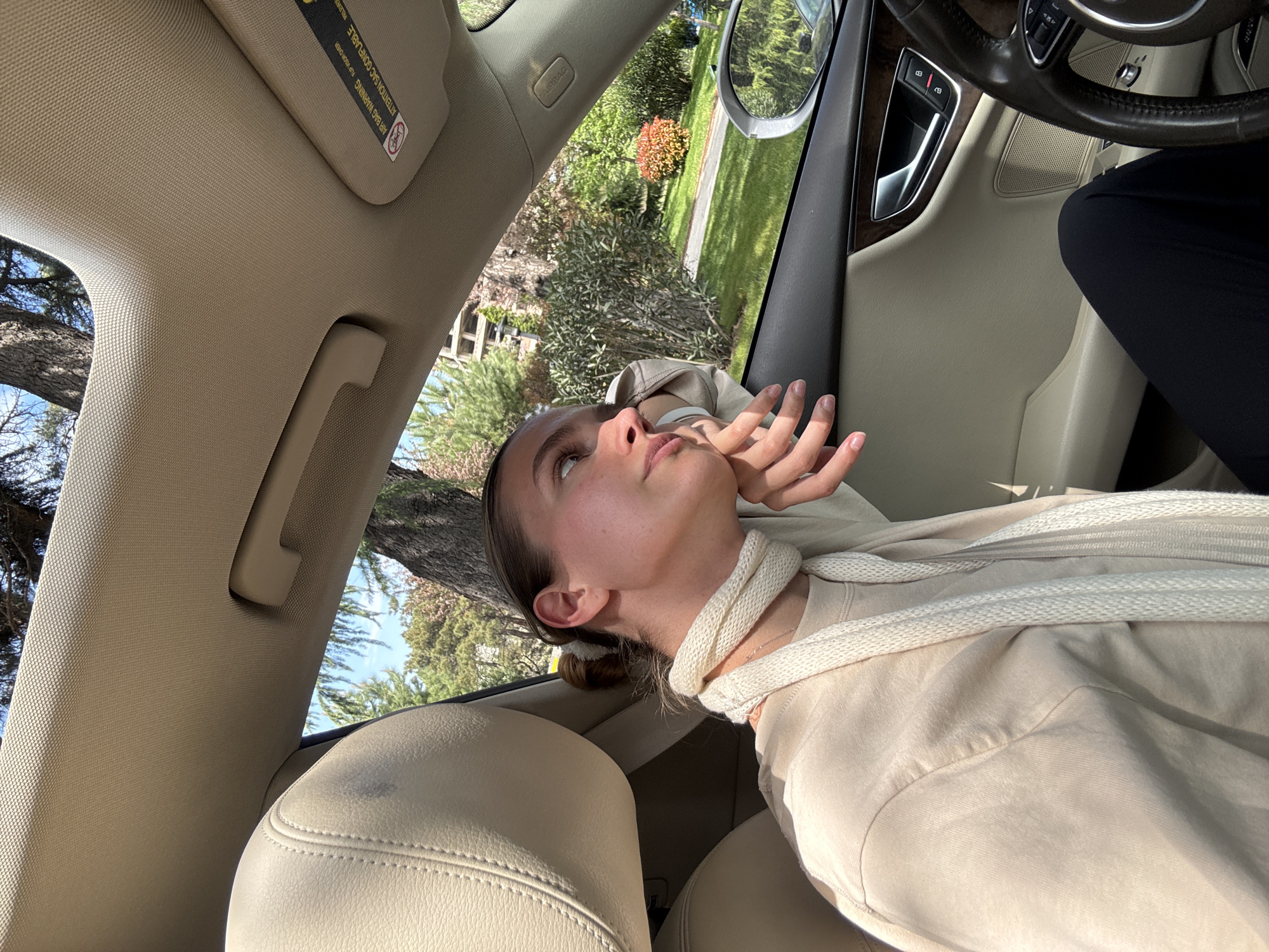 A woman is sitting in the passenger seat of a car. She is fair-skinned with brown hair pulled back in a bun and is wearing a light-colored sweatshirt and a rope-like scarf. Her head is tilted back, and she is looking upwards with her hand near her face. The interior of the car is beige and includes the passenger seat, door, steering wheel, and sun visor. Outside the car window, there is greenery that includes trees, bushes, and grass. The location is not discernable as Tbilisi, Georgia.