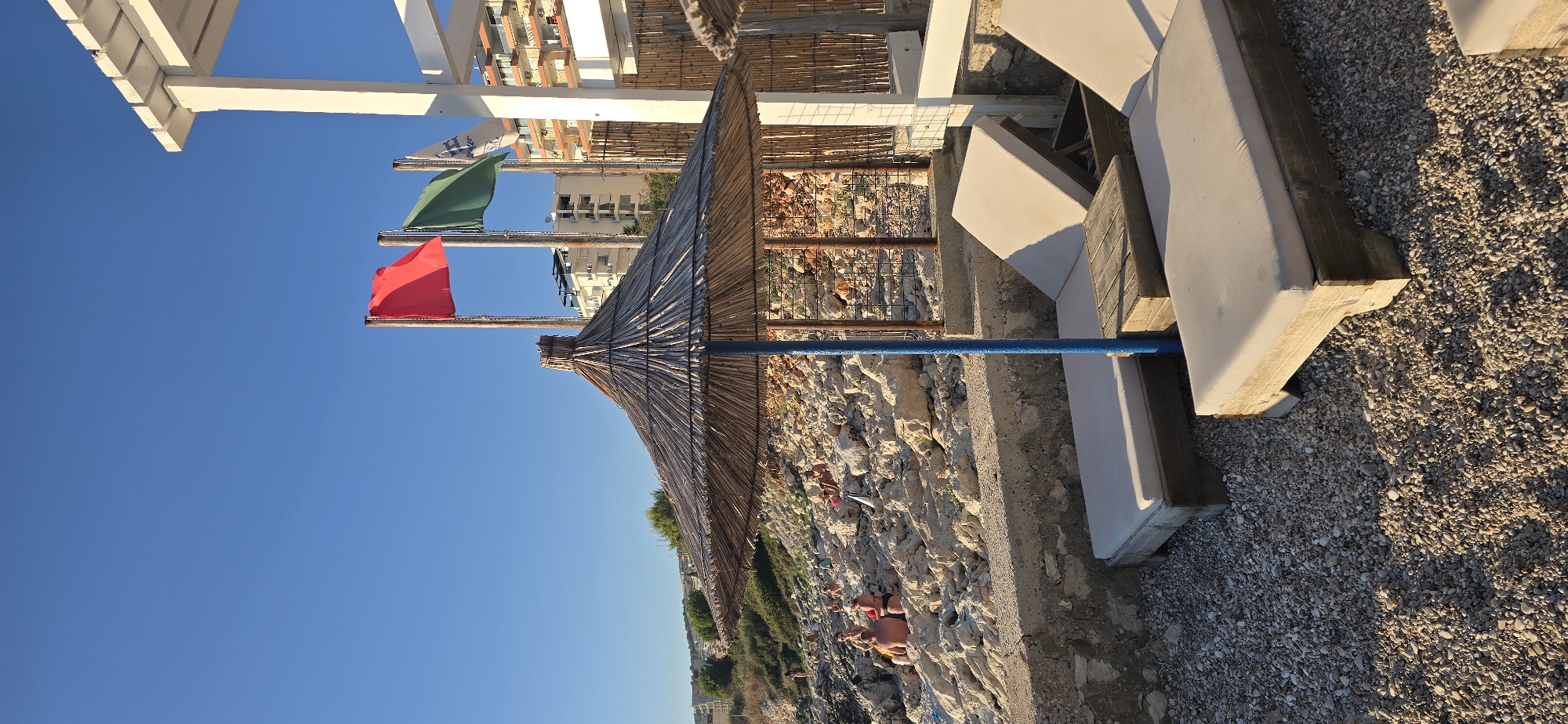 This is a vertical, eye-level shot of a beach in Saranda, Albania. In the foreground, two empty lounge chairs with white cushions are set up on a gravelly beach. A straw umbrella provides shade over one of the chairs, with its blue pole visible. A small wooden table sits between the two chairs.
Behind the chairs, a low stone wall separates the gravel beach from a rocky incline. Several people are scattered on the rocks, some sitting, some standing, engaged in various leisure activities like sunbathing and talking. The rocky terrain is dotted with sparse vegetation.
Further in the background, a white structure, possibly a lifeguard stand or part of a beach facility, is visible. Three flags are flying from poles: a red flag on the left, a green flag in the middle, and a white flag with blue markings on the right. Beyond the beach facilities, modern apartment buildings with balconies rise up on a hillside, indicating the urban proximity to this beach. The sky is a clear, bright blue, suggesting a sunny day.