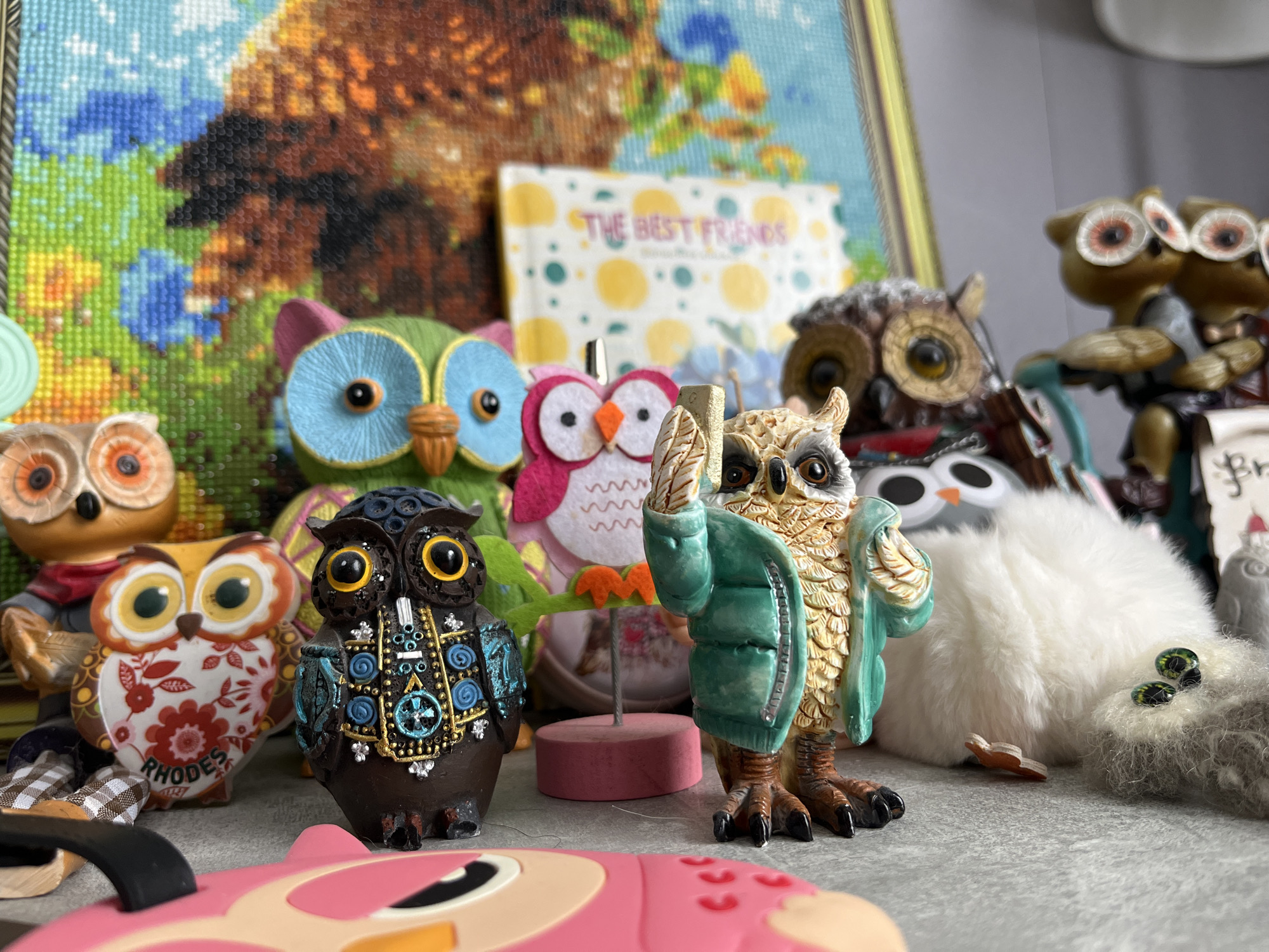 The image shows a collection of owl figurines. These owls are of varying sizes, colors, and materials, including ceramic, felt, and possibly resin. The arrangement is a cluttered display on a flat surface, which appears to be a countertop or table. In the background, there is a framed piece of art depicting an owl, as well as a book titled "The Best Friends." The figurines are densely packed together. A figurine labeled "RHODES" is visible.
There is no information in the image that provides evidence about the location in the city of Shushary, Russia.