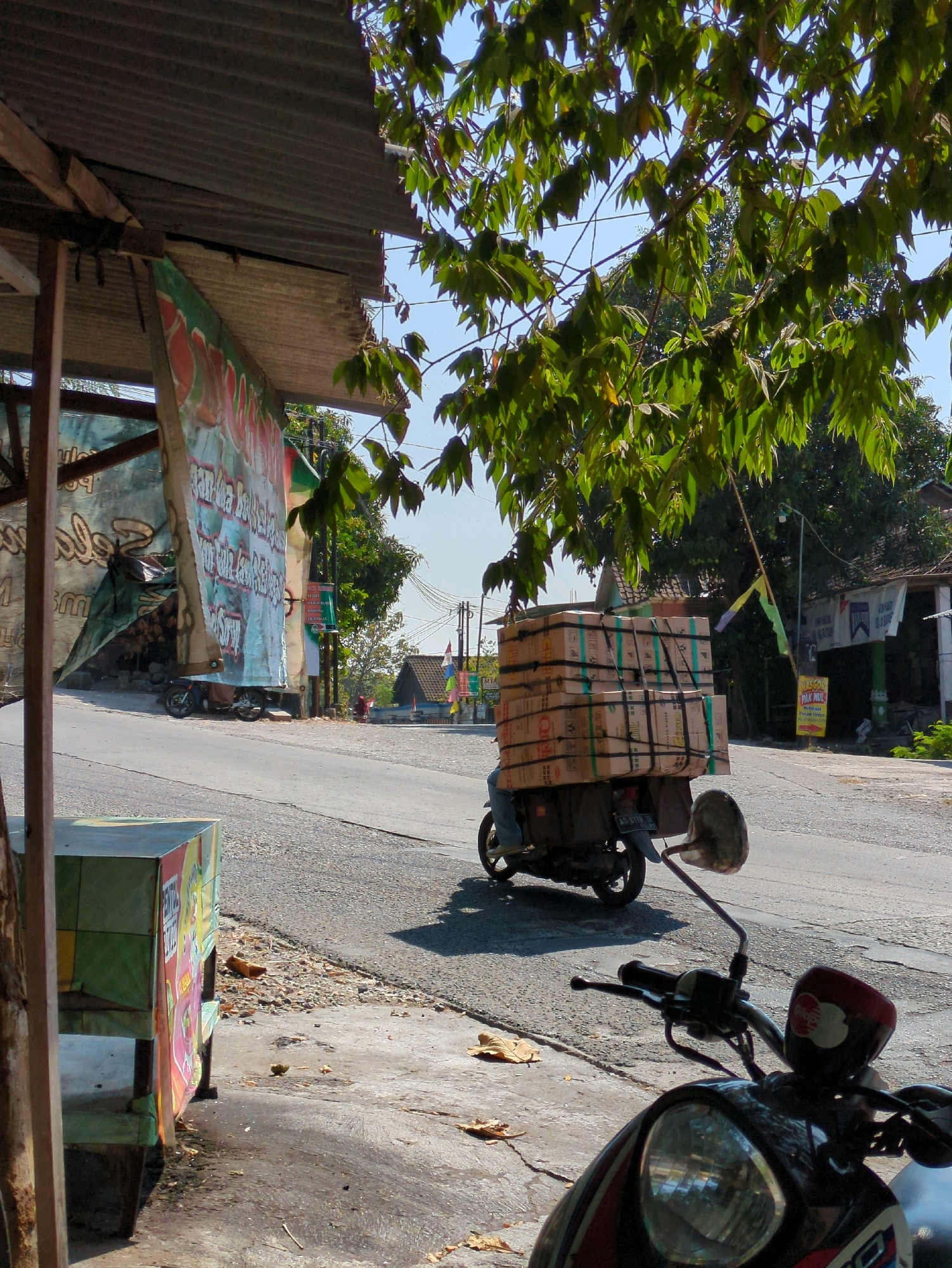 In Sindon, Indonesia, a scooter laden with a towering stack of cardboard boxes is traveling down a paved road. The boxes are secured with black straps. The scooter is on the right side of the road, moving away from the camera. Sunlight filters through the leaves of a tree in the foreground, casting shadows on the road and a colorful, weathered display case to the left. A large, ripped tarp hangs from a makeshift shelter to the left of the road, revealing a motorcycle parked behind it. Buildings and utility poles are visible in the background. The scene appears to be an outdoor commercial or residential area.