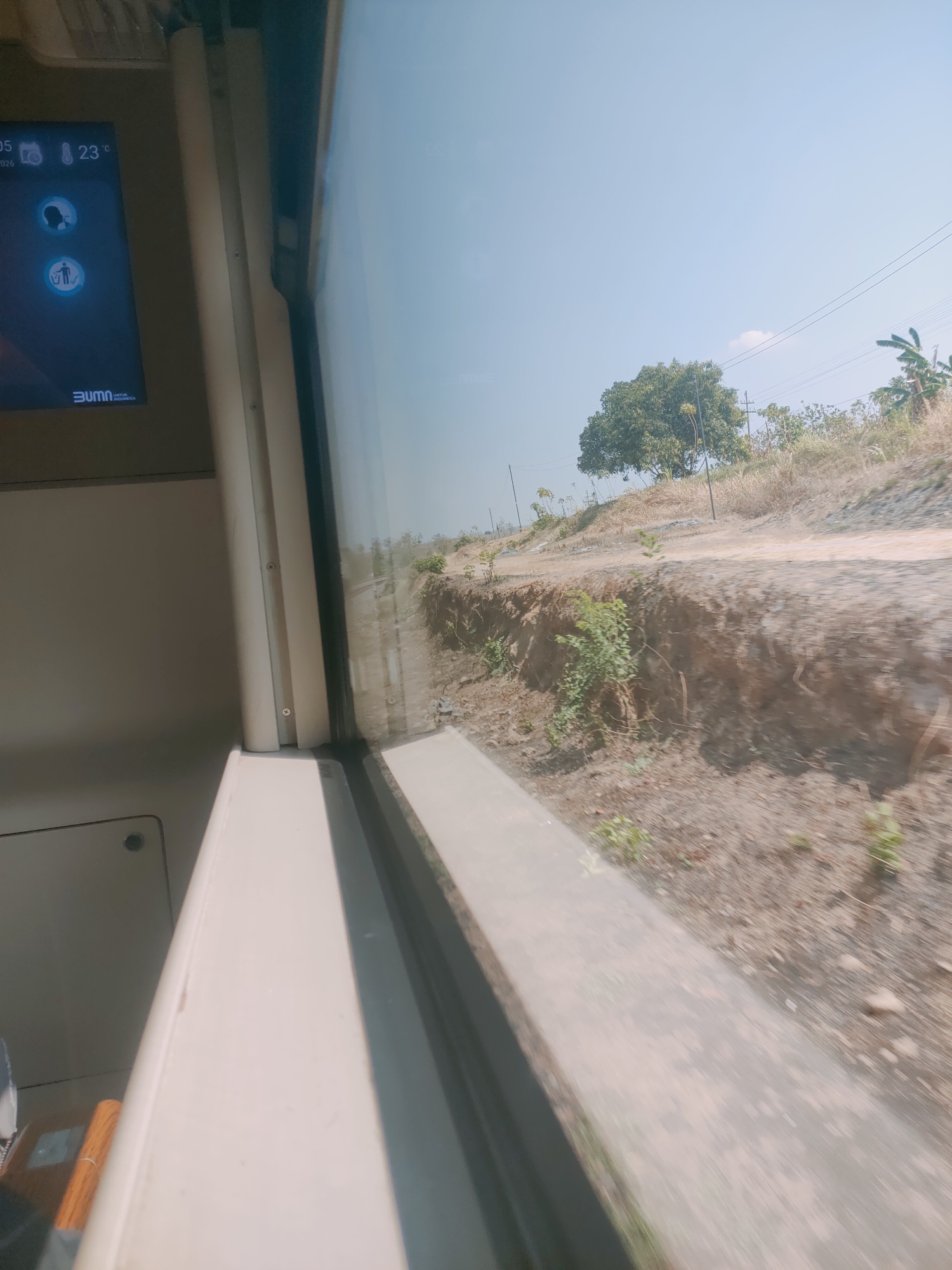 The image depicts a view from inside a moving vehicle, likely a train, looking out a window. The foreground shows a portion of the vehicle's interior, including a beige-colored panel and the edge of a seat with orange-wrapped items. The window reveals a landscape with dry, dusty terrain, a dirt road, a steep embankment with sparse vegetation, and a single large tree. Power lines are visible against a clear blue sky. A digital display screen on the left side of the interior shows "05" and "23°C", along with icons and the "BUMN UNTUK INDONESIA" logo. The scene is captured during the day, likely in a rural or semi-rural area of Wilangan, Indonesia, suggested by the "BUMN UNTUK INDONESIA" text.