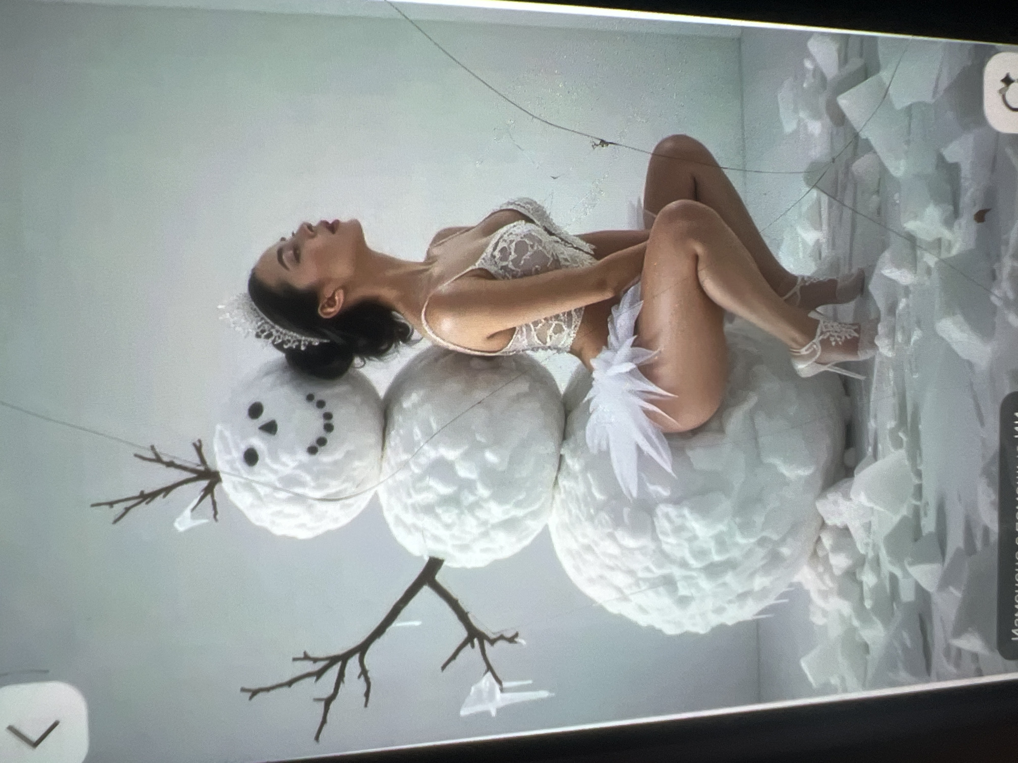This is a studio photograph. A woman wearing a white lace bralette and a fluffy white skirt is posed next to a three-tiered snowman. The snowman has twig arms, a carrot nose (not visible), and a smiling face made of dark dots. The woman is wearing a tiara and white high-heeled shoes with lace accents. The ground around the snowman and woman is covered in broken pieces of ice or snow. The background is a plain, light-colored wall. There is no indication of the location being Kara-Dıykan, Kyrgyzstan; it appears to be a set designed for a photoshoot.