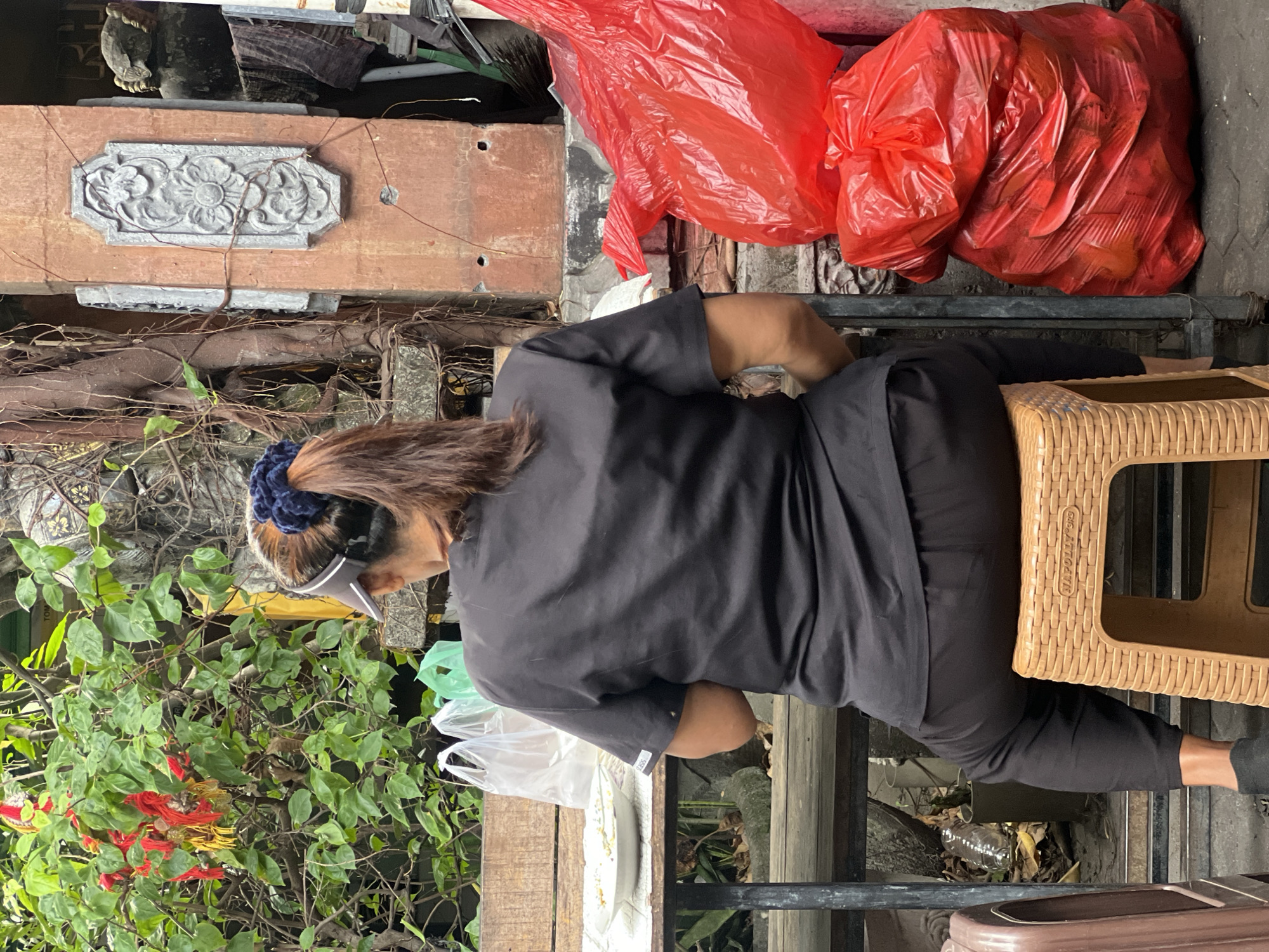A person wearing a black scrub top and black pants, with their back to the camera, is sitting on a light brown wicker stool. They have brown hair pulled back in a ponytail with a dark blue scrunchie and are wearing a clear visor. In front of them, on a wooden table, is a white plate with some food and several plastic bags. To their right, a stack of two bright red plastic bags are visible, one on top of the other. The background shows a rough, textured wall with some decorative stone carving, overgrown vines, and lush green foliage with red flowers. The scene appears to be outdoors, possibly in a courtyard or alleyway in Kuta, Indonesia, given the building materials and vegetation.