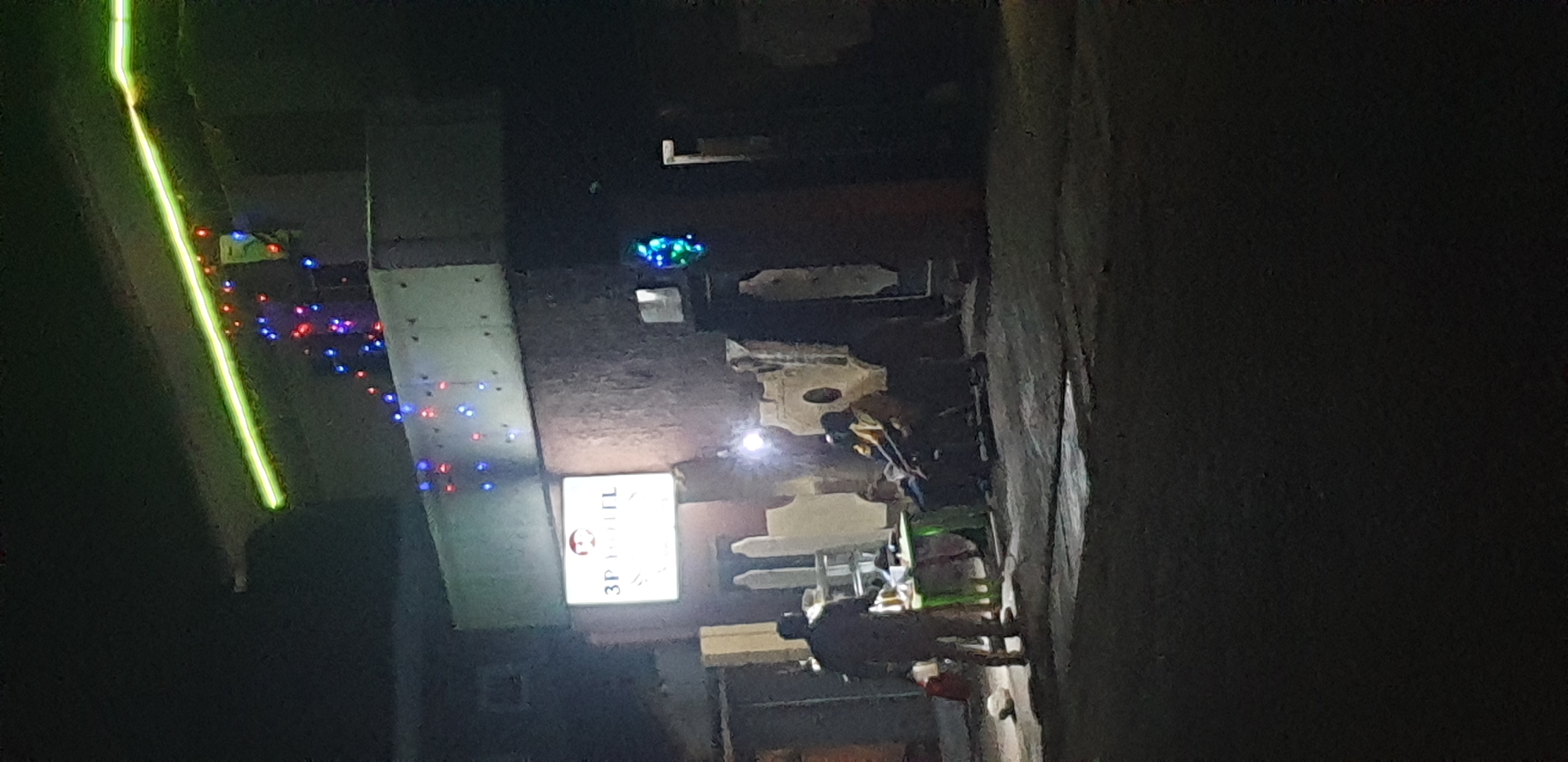 The image depicts a nighttime scene in Narayi, Nigeria, featuring the exterior of a building identified by an illuminated sign as "3P HOTEL."
The building's upper facade features a horizontal, bright green linear light strip. Below this, on a darker, textured section of the building, numerous small, multi-colored string lights, primarily red and blue, are affixed. A rectangular, internally lit sign on the building's front displays "3P HOTEL" in red lettering with a circular red logo above it. Further to the right on the building, another cluster of blue and green lights is visible.
At ground level, there are several individuals and objects. One person, seen from behind, stands on the left side of the frame near a small, green-topped table. Several items are on the table, but their specifics are indistinct. Behind this person, a white, multi-tiered stand or shelf is visible. Further into the scene, to the right of the standing person, two or three individuals are seated on a bench or chairs, appearing to interact. Two of these individuals are wearing light-colored, possibly yellow, shirts. A bright, localized light source illuminates the area around the seated individuals and the table.
The ground surface is dark and uneven, possibly concrete or hardened earth, with some lighter areas reflecting light. Various irregularly shaped, light-colored objects, possibly concrete blocks or decorative elements, are present in the mid-ground. The overall scene is dimly lit by artificial sources against a dark night sky.