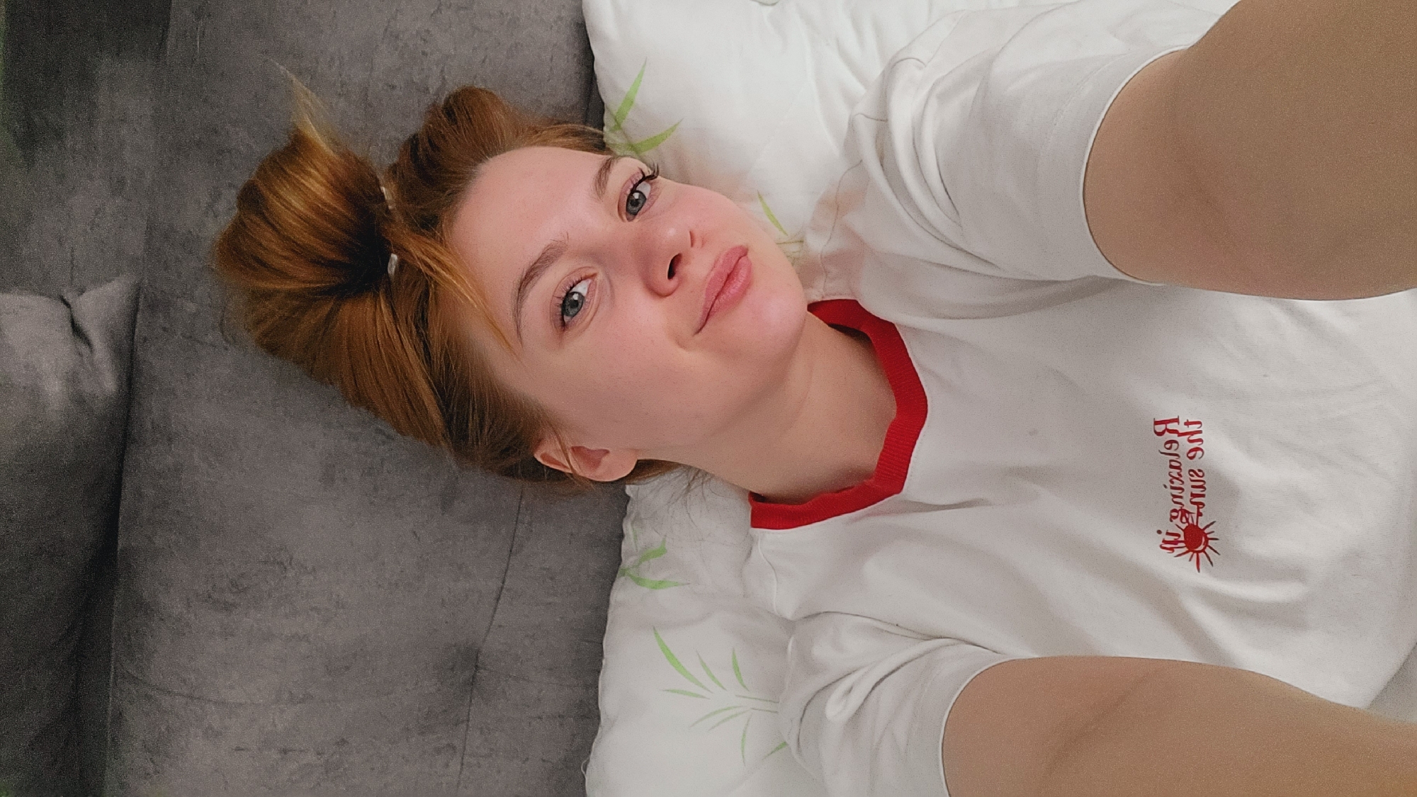A woman lies on a gray couch or sofa, her head resting on a white pillow with a green bamboo pattern. She is facing the camera, her head slightly tilted. Her hair is reddish-brown and styled in a bun. She wears a white t-shirt with a red collar and the text "Raising Sun" printed in red. The location is Chaikovsky, Russia.