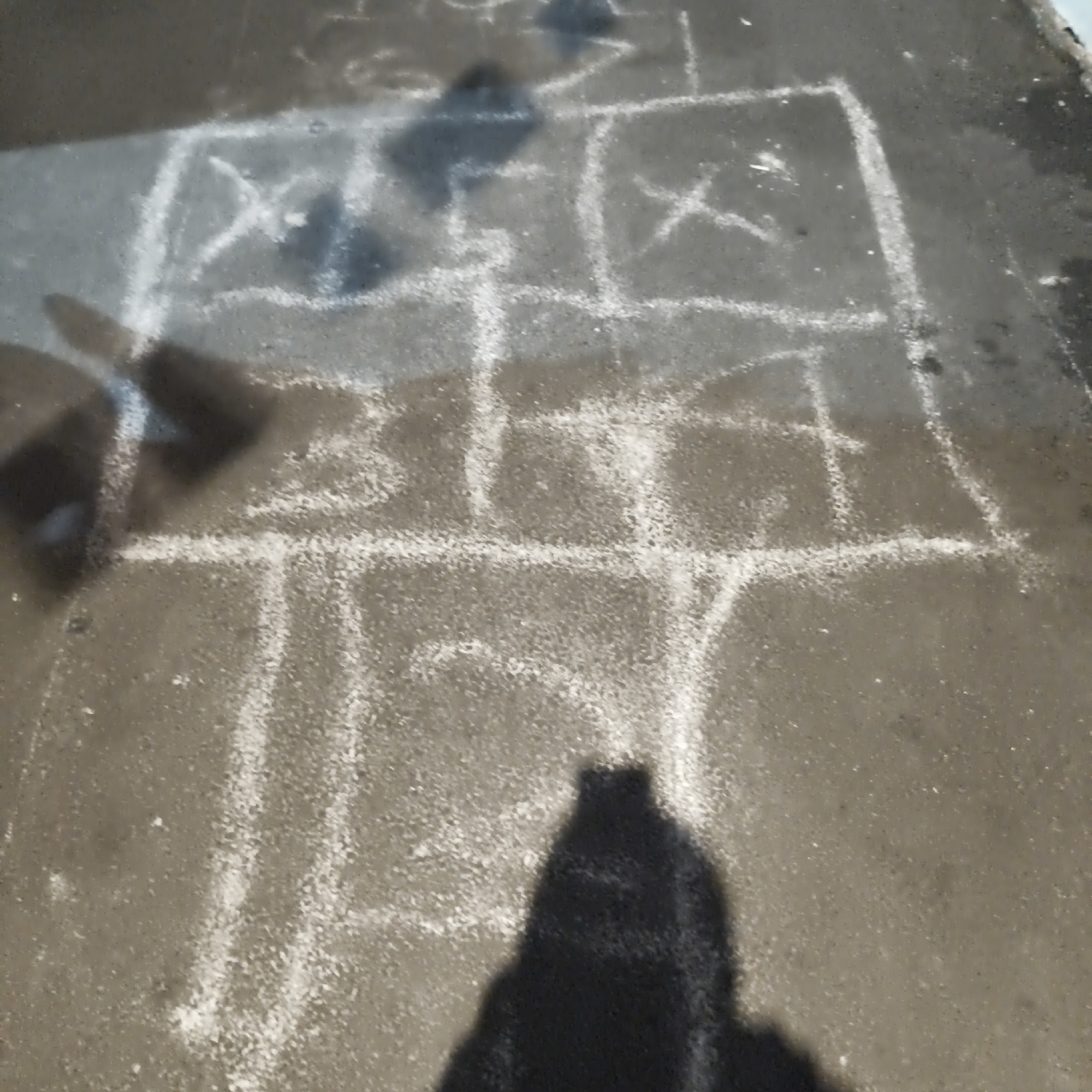The image captures a hopscotch grid drawn with white chalk on a wet, paved surface, likely a sidewalk or road in the city of South Jakarta, Indonesia. The grid is composed of squares and rectangles, with some squares containing chalk marks that resemble "X"s. A dark shadow, presumably cast by a person, is visible in the foreground, obscuring a portion of the hopscotch grid. The wet pavement suggests recent rainfall or damp conditions. The overall scene is outdoors, and the chalk markings indicate a recreational activity.