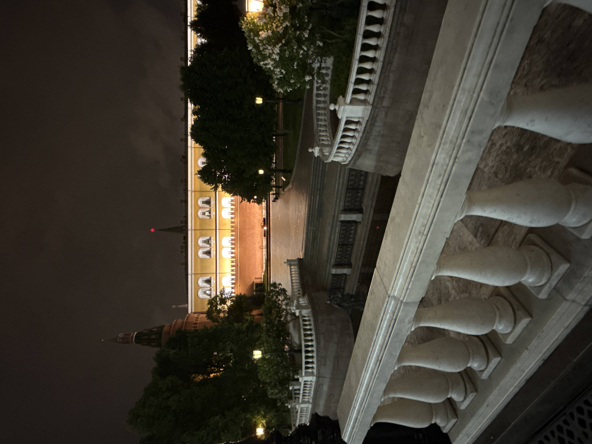 The image shows a night view of the Moscow Kremlin, as seen from a perspective behind a white stone balustrade. In the background, the yellow facade of the Kremlin building is illuminated, with windows visible in a repetitive pattern. A red star is visible atop one of the towers. The view also includes a darker, more textured tower, shrouded in shadow and foliage. A walkway lit by lamps stretches between the building and the viewer's position. Trees and greenery are present, some with white blossoms, adding depth to the scene. The foreground is dominated by the white balustrade, featuring classic architectural details.
