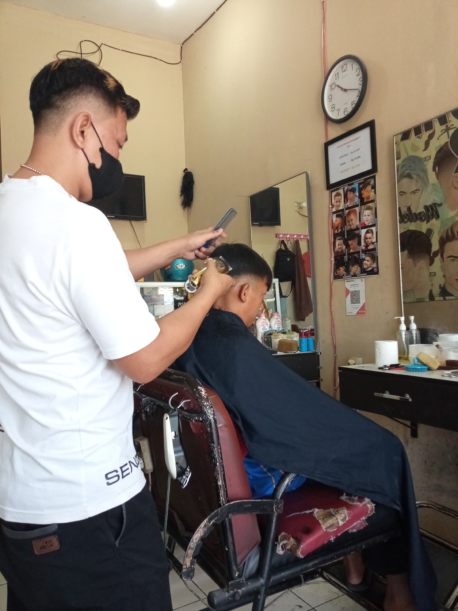 In a modest barbershop in Sariwangi, Indonesia, a barber, wearing a white t-shirt and black mask, is cutting the hair of a young male client. The client is seated in a worn, red leather barber chair, draped in a black cape. The barber is using a hair clipper and a comb, actively styling the client's hair. The barbershop's walls are a neutral beige, adorned with a clock, a price list for haircuts, and posters featuring various hairstyles. A mirror on the wall reflects a portion of the room, revealing shelves stocked with hygiene products and barber tools. The overall atmosphere suggests a typical local barbershop with a functional, no-frills setup.
