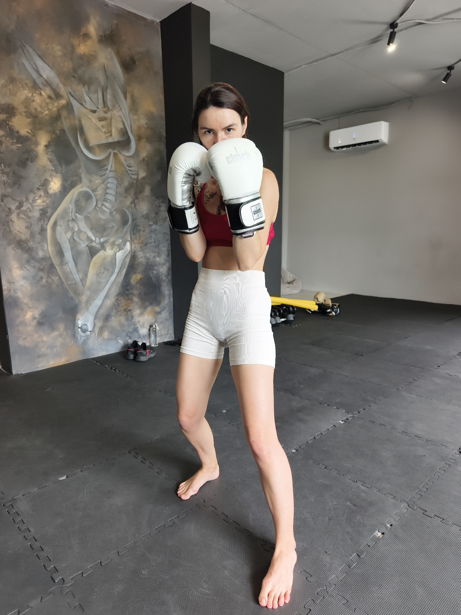 This full-shot image captures a woman in a boxing stance within an indoor gym setting, likely located in Tyumen, Russia. She stands barefoot on a dark grey interlocking rubber mat floor, directly facing the camera with a focused and serious expression.
The woman, appearing to be in her 20s or 30s, wears a red sports bra or crop top and white ribbed athletic shorts. Her hands are held up in a defensive position, encased in white boxing gloves that prominently display the brand name "Clinch" in black text on the back of each hand. Her dark hair is pulled back, and her gaze is direct and intense.
The background behind her left side features a large, abstract mural on a dark grey wall. This artwork depicts skeletal or anatomical forms in various shades of grey and beige, offering a unique artistic element to the gym. Below the mural, a pair of black athletic shoes and a clear water bottle are visible on the floor. On her right side, the background includes a plain grey wall with a white air conditioning unit mounted high up. The ceiling is also grey, equipped with black track lighting fixtures that illuminate the space. Further in the background on her right, some yellow fitness equipment and other dark objects can be seen on the mats, suggesting various training tools.