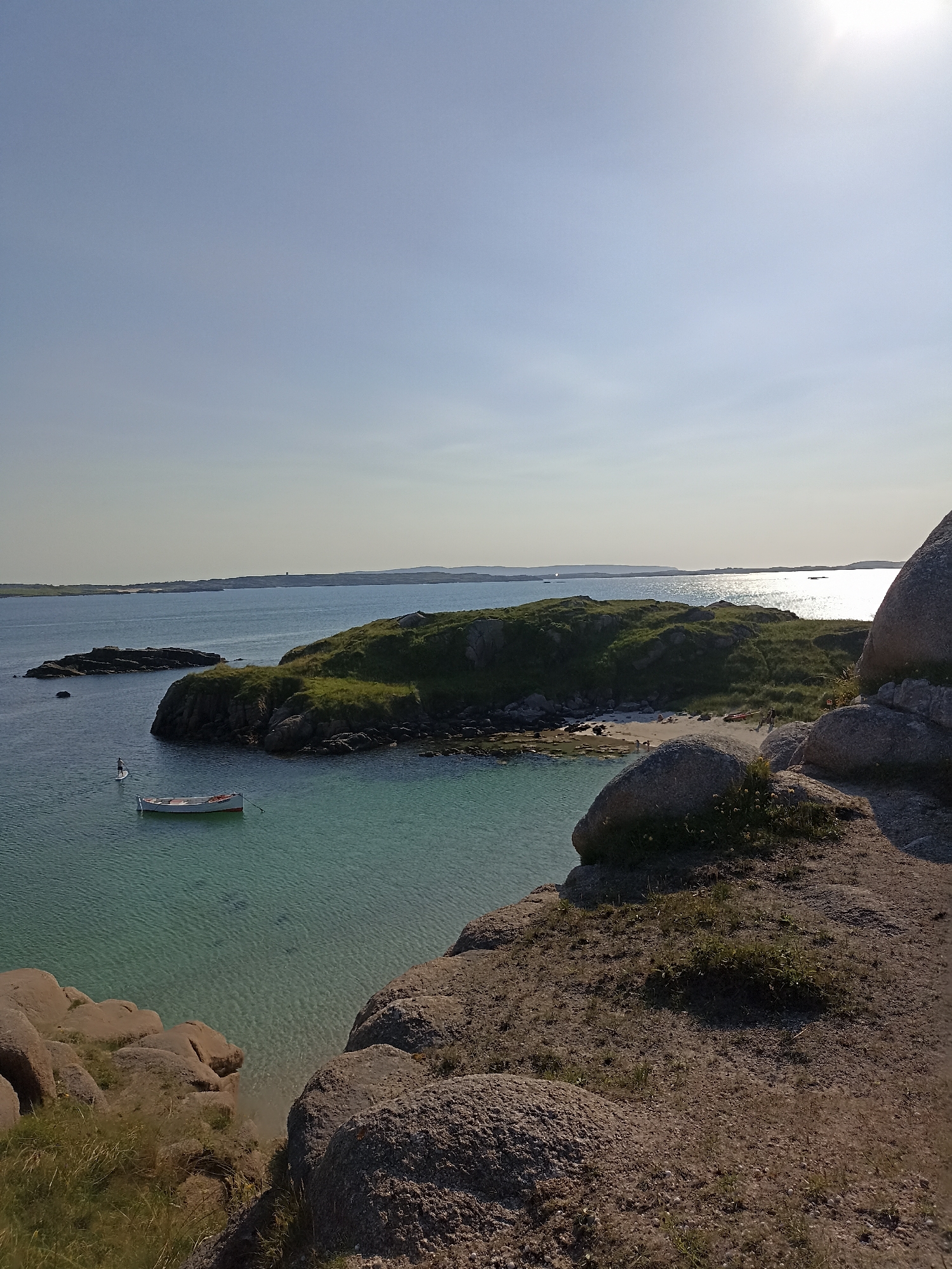 The image depicts a serene coastal landscape. In the foreground, large, rough granite boulders are prominent, with sparse grass growing between them. A clear, turquoise body of water occupies the middle ground, revealing the sandy seabed below. A small, white boat with orange trim floats on the water, anchored by a rope. Further in the distance, a green, grassy island with rocky outcrops rises from the sea. A single person on a paddleboard is visible in the water between the island and the foreground. The background shows a calm sea stretching to the horizon, with distant islands or landmasses under a clear, bright sky. The sun is high and slightly to the right, casting a bright glare on the water. The overall impression is one of a peaceful, natural setting on a sunny day. The location is identified as Carnboy, Ireland.