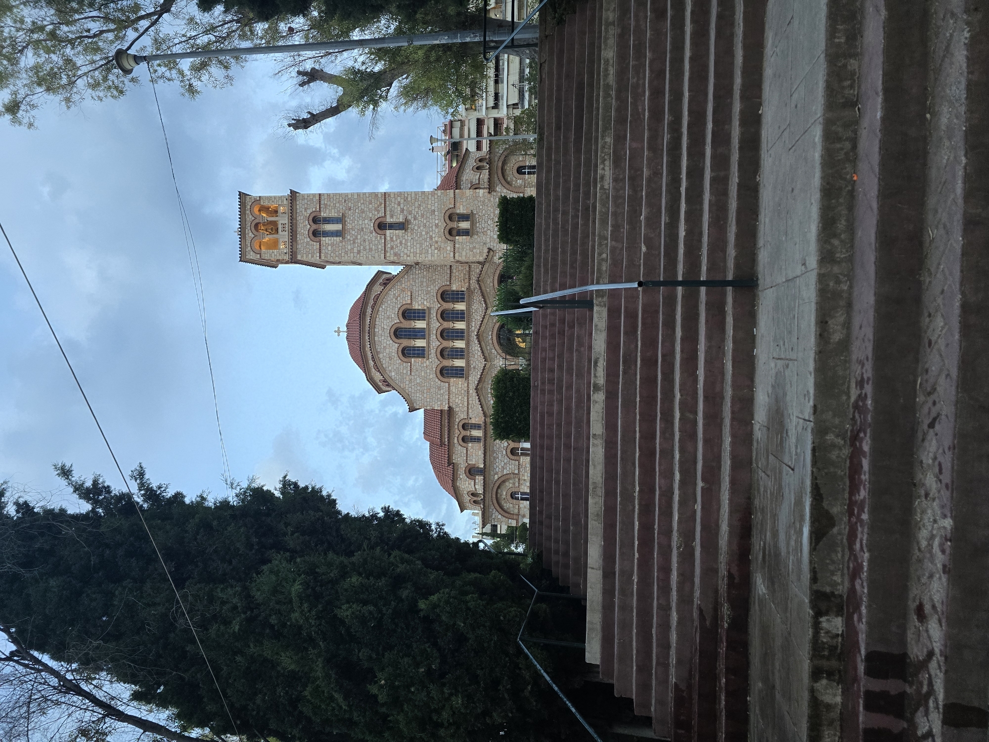 The image shows a stone church with a bell tower and a dome, likely in Nea Ionia, Greece. The church is constructed from light-colored stones and features arched windows. The bell tower has open archways at the top, revealing illuminated interiors. A red-tiled roof covers the dome and parts of the church.
In the foreground, a wide staircase made of stone leads upwards. A metal handrail runs along the side of the stairs. Dense evergreen trees frame the scene on the left, with some bare branches visible in the upper left corner. A lamppost is also visible above the trees, with power lines stretching across the sky.
The sky is overcast with some lighter patches, suggesting it is either early morning or late afternoon. The lighting is diffused, and the scene appears to be quiet and peaceful. There are no people visible in the image. The overall impression is one of a serene, historical religious site. No visible text is present.