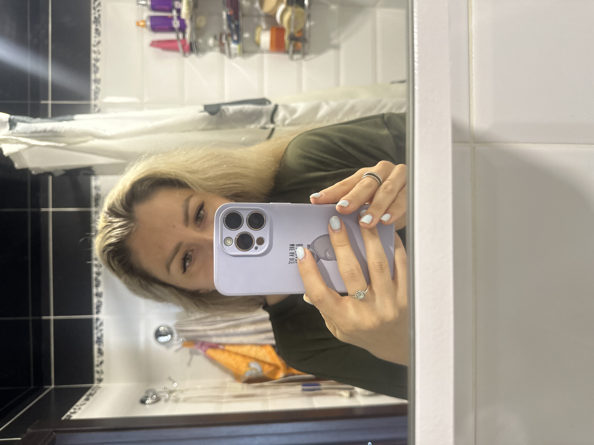 In a bathroom located in Novoaltaysk, Russia, a woman with blonde hair takes a selfie in a mirror. She is holding a lavender phone case with a phone inside, capturing her reflection. The phone case has writing on it in Russian that reads "МОЙ ВЫБОР ВЕ" and a circular graphic. She is wearing a dark green shirt, and her nails are painted a light blue. Two rings are visible on her left hand. The background shows a tiled wall with black and white accents, a shower curtain, and shelves with various toiletries.