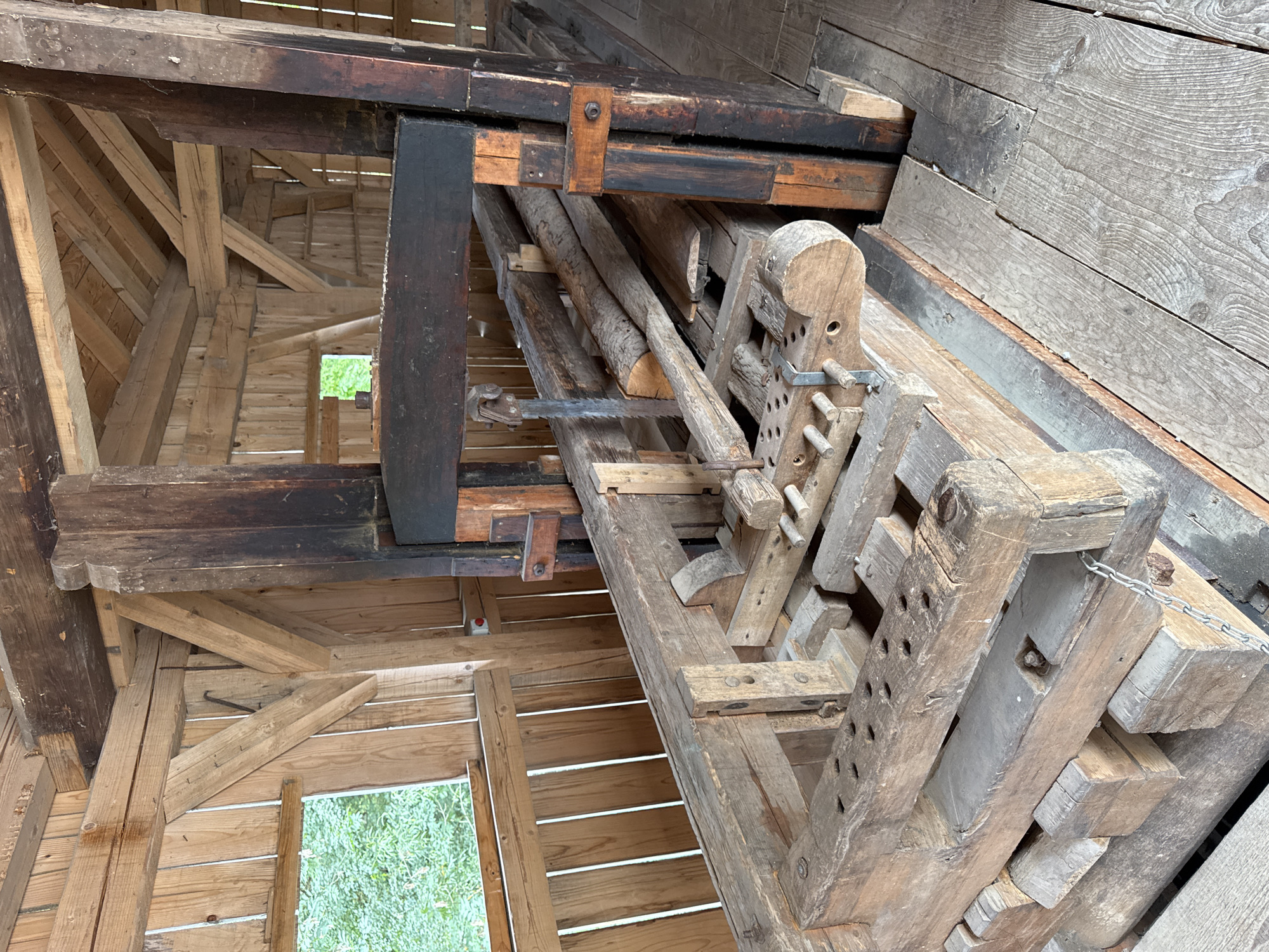 The image shows the interior of a historic wooden sawmilling establishment, likely located in or near Postojna, Slovenia, given the context of the provided information. The scene is dominated by a large, antiquated wooden saw mechanism. This machinery is constructed from heavy, aged timbers, showing signs of wear and tear. A vertical saw blade is visible, suspended by a mechanism above a wooden track system. Logs and planks are positioned on this track, awaiting or having undergone the sawing process. The structure itself is built of rough-hewn lumber, with exposed beams and wooden walls. A small window with a view of green foliage is present in the background. The overall impression is of a rustic, functional space dedicated to traditional woodworking.