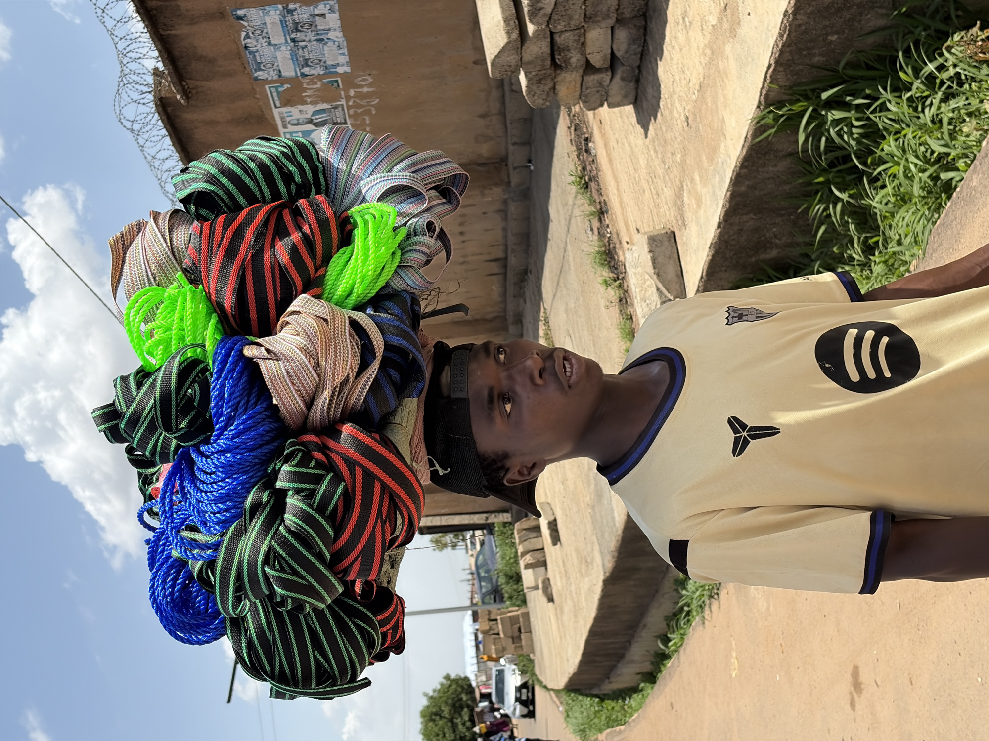 The image captures a vibrant street scene in Gura Top, Nigeria, on a bright, sunny day with a clear blue sky and scattered white clouds. The main subject is a young man, likely a vendor or hawker, expertly balancing a large, colorful bundle of ropes or straps on his head.
The bundle consists of various distinct colors and patterns, including bright neon green, royal blue, black with red stripes, black with green stripes, and patterned beige. The man wears a light-colored (possibly cream or light yellow) short-sleeved t-shirt with dark blue and black trim on the collar and sleeves. His shirt prominently displays two black logos: the Spotify logo (three curved lines) on the right side of his chest, and a stylized three-pronged symbol (possibly an athletic brand logo like a stylized 'Y') on the upper left chest. He also wears a black baseball cap backward. The man is looking upwards and slightly to his right with a neutral expression, his mouth slightly open.
In the background, a concrete structure, possibly a wall or the underside of a bridge, features barbed wire along its top edge. Fragments of old posters and flyers are visible on this structure, with the numbers "53270" partially legible on one piece of paper. Lush green foliage grows along the base of the concrete structure on the right side. In the mid-ground, a bustling street with blurred vehicles and people suggests an active urban environment. The strong shadows cast by the sun indicate it is likely mid-day or early afternoon.