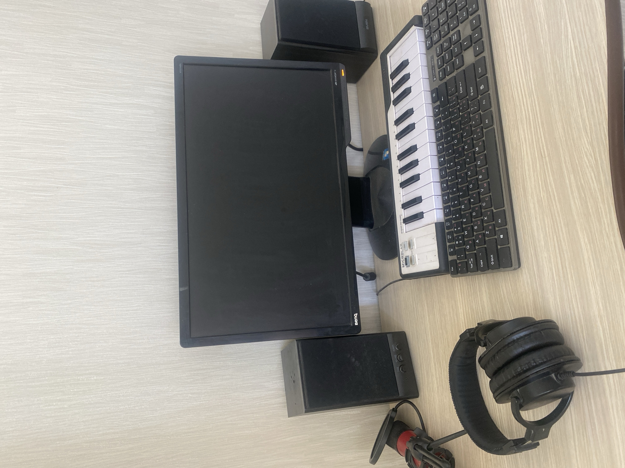 The image shows a desk with a computer monitor, two speakers, a keyboard, a MIDI controller, headphones, and a microphone. The computer monitor is black and has a BenQ logo on the bottom bezel. The speakers are black and rectangular. The keyboard is black and appears to be a standard QWERTY keyboard with some additional keys. The MIDI controller is white and has several keys and buttons. The headphones are black and over-ear. The microphone is red and black and is attached to a stand. The desk is made of wood. The background is a textured wall. The scene is a workspace, likely for music production or audio recording. There are no people present in the image. The location in Chișinău, Moldova is not discernible from the image.