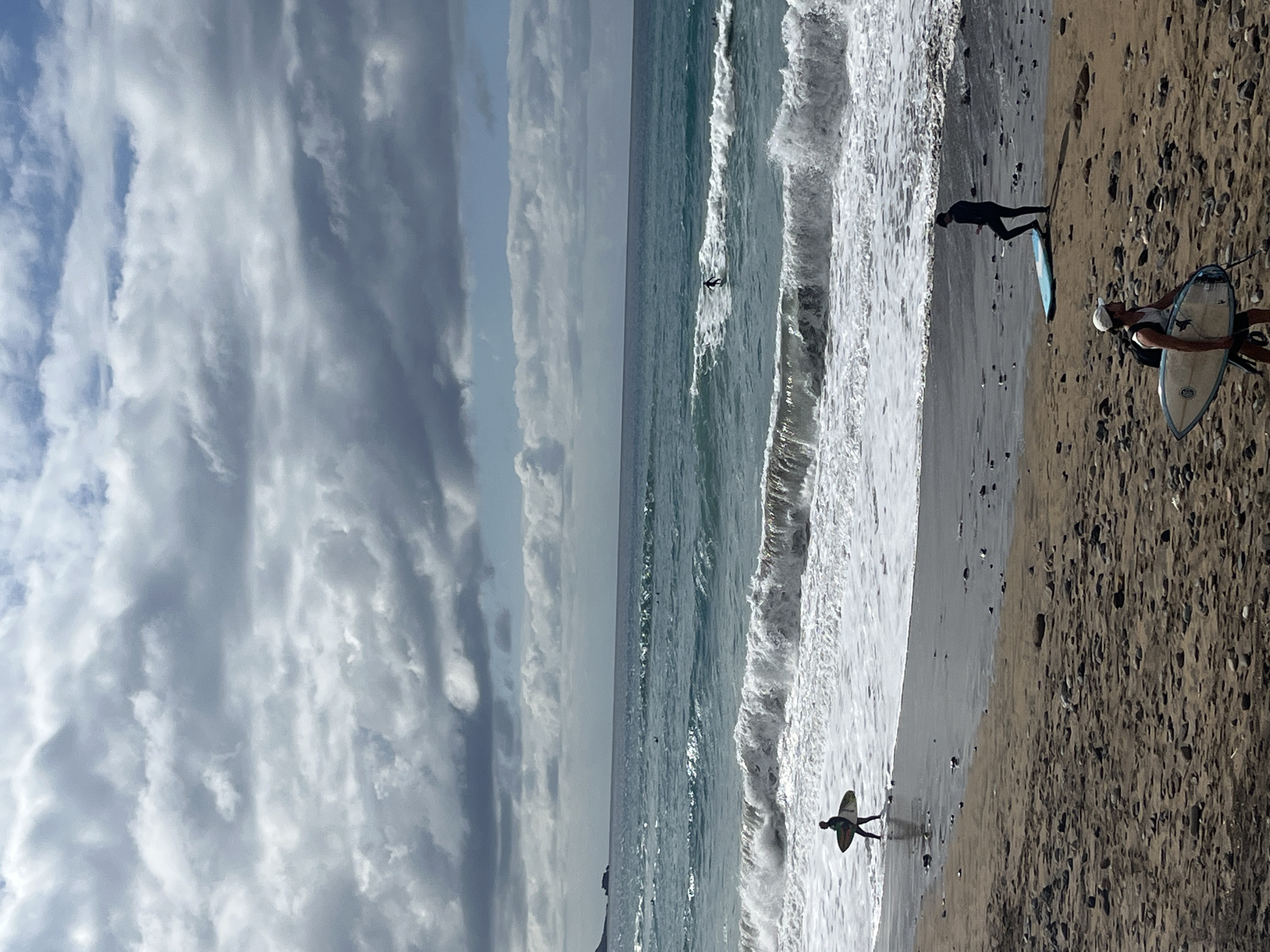 The image captures a dynamic beach scene, likely in Las Palmas, Spain, based on the provided context. The perspective is broad, encompassing the sky, ocean, and sandy shore.
The sky, occupying the left portion of the image, displays a mix of blue patches and extensive white and grey clouds, some appearing wispy and elongated. Below the sky, the ocean stretches across the middle of the frame, its waters a deep blue-green. Several waves are breaking, creating lines of white foam that extend towards the shore. In the deeper water, small dark figures, presumably other surfers, are visible as specks, with one person in a dark wetsuit actively riding a wave in the mid-ground.
The right side of the image features a sandy beach. The sand appears wet closer to the water's edge, transitioning to a drier, speckled texture further up, scattered with small dark pebbles.
There are multiple subjects on the beach and in the water. On the dry sand in the foreground, a person wearing a white cap and dark top is seen holding a surfboard with a white deck and blue bottom. Closer to the water, another individual in a dark wetsuit stands in the shallow surf, with a light blue surfboard lying on the wet sand beside them. Further down the beach, another person in a dark wetsuit is positioned in the water with their surfboard.
The activity taking place is surfing. The individuals are either preparing to enter the water, waiting for waves, or actively riding them. The scene suggests a popular surfing spot on a day with moderate waves and a partly cloudy sky. No discernible text is visible in the image. The time of day appears to be daytime, possibly late morning or early afternoon, given the bright yet diffused light.