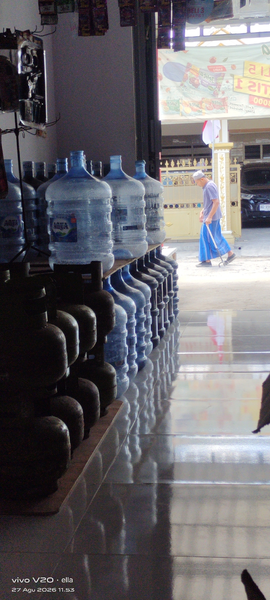 The image depicts an outdoor scene in Simo, Indonesia, likely a storefront or market area. In the foreground, a row of large, clear plastic water jugs filled with water is stacked on shelves. Behind these jugs, a person wearing a light blue robe and a white cap walks with a cane. The person is walking towards the right side of the frame, away from the viewer. In the background, there is a building with a decorative fence and a parked car. A banner with text and graphics is visible hanging from the building. The ground is paved and reflective, showing distorted reflections of the jugs and the surroundings. The overall lighting suggests it is daytime, with sunlight casting shadows. The image is watermarked with "vivo V20 · ella" and "27 Agu 2026 11.53".