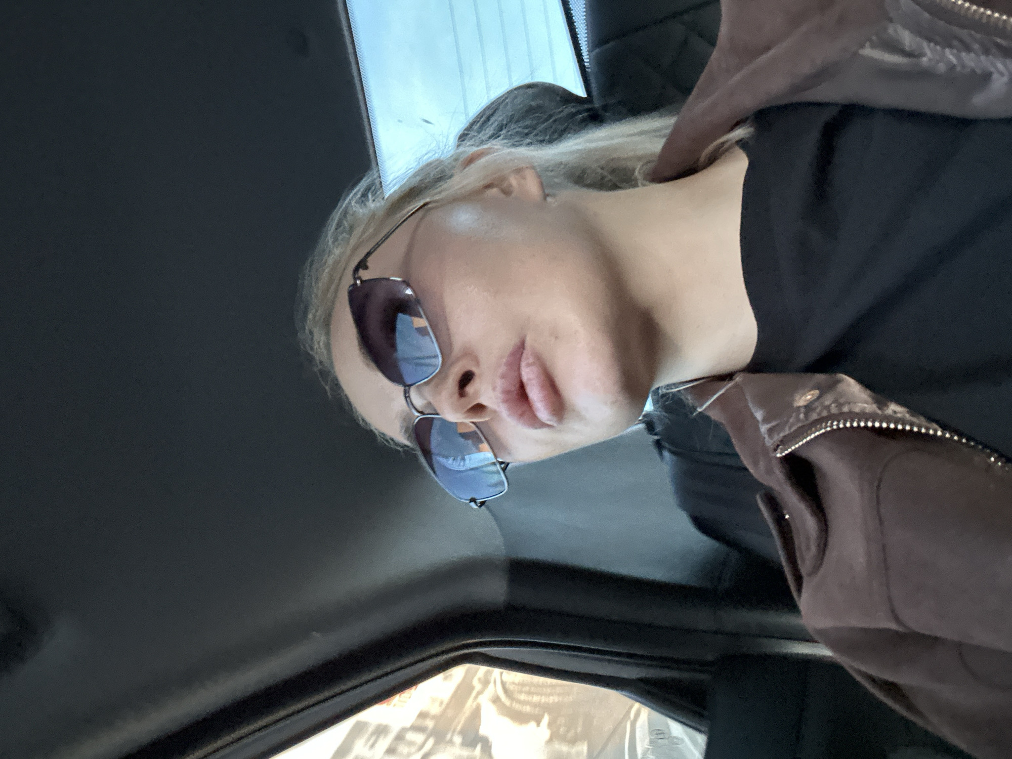 A woman with blonde hair and sunglasses is positioned inside a car. The interior of the vehicle is visible, including the dashboard and part of the side window. The woman wears a dark shirt and a brown jacket. The location is the city of Megion, Russia.