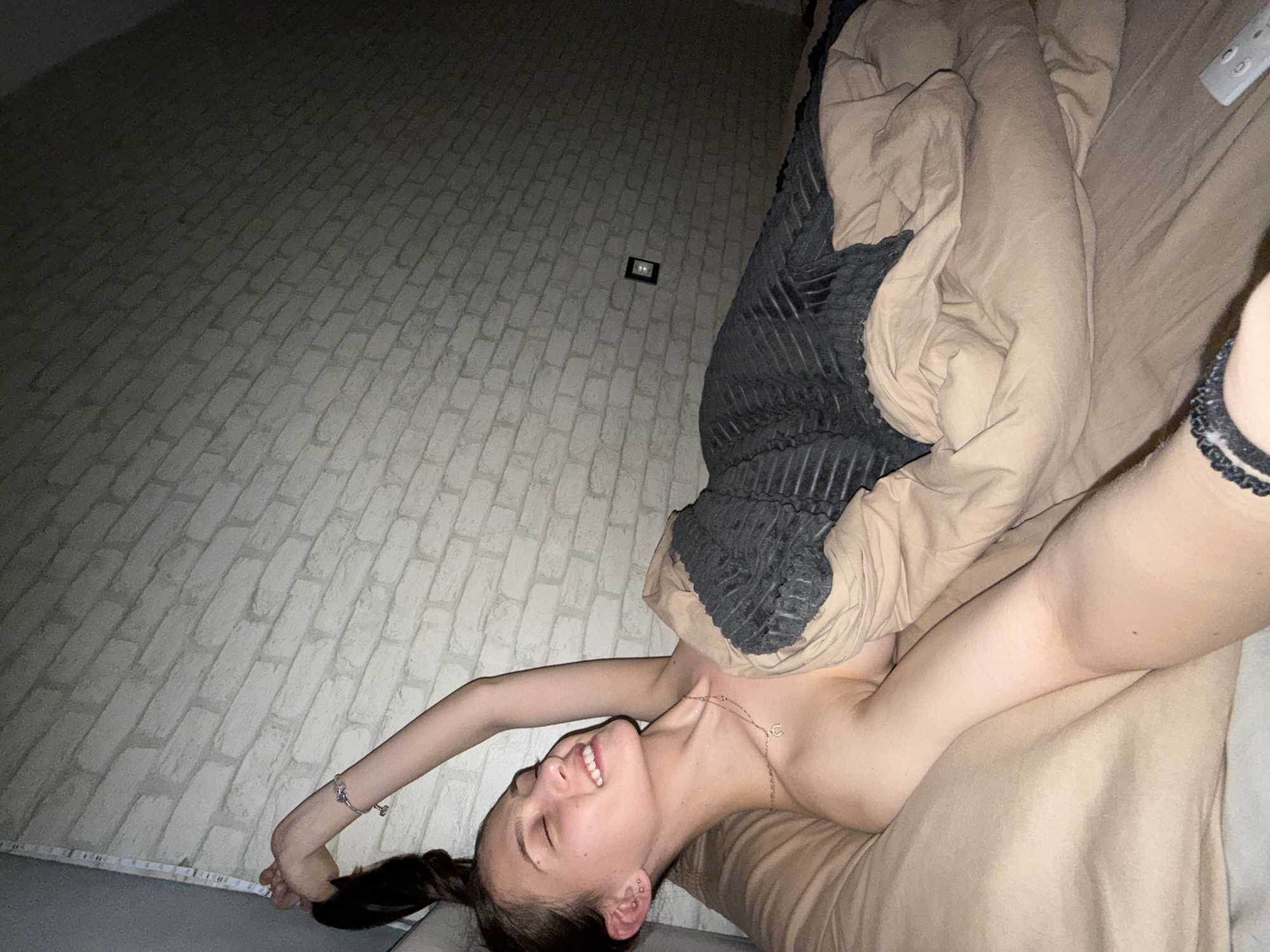 The image displays a young woman lying on a bed or sofa, captured from an overhead perspective that is rotated. She is smiling broadly with her eyes closed, conveying a sense of happiness and relaxation. Her dark hair is pulled back, and she has visible ear piercings and a delicate gold chain necklace with a "C" pendant. Her arms are extended upwards, stretched out comfortably.
She is partially covered by a beige duvet or blanket, with a dark grey or black textured blanket also draped nearby. The setting is an indoor room, characterized by a light-colored wall featuring a distinct brick-like pattern. The lighting in the room is dim, suggesting it is evening or night.
A small black rectangular device with two white lights is mounted on the brick wall. On the bed, near her head, a white remote control is visible. Along the bottom edge of the brick wall, there's a white strip that appears to be either a measuring tape or an LED light strip. The overall mood is one of peace and contentment. The location context for this scene is Sofia, Bulgaria.