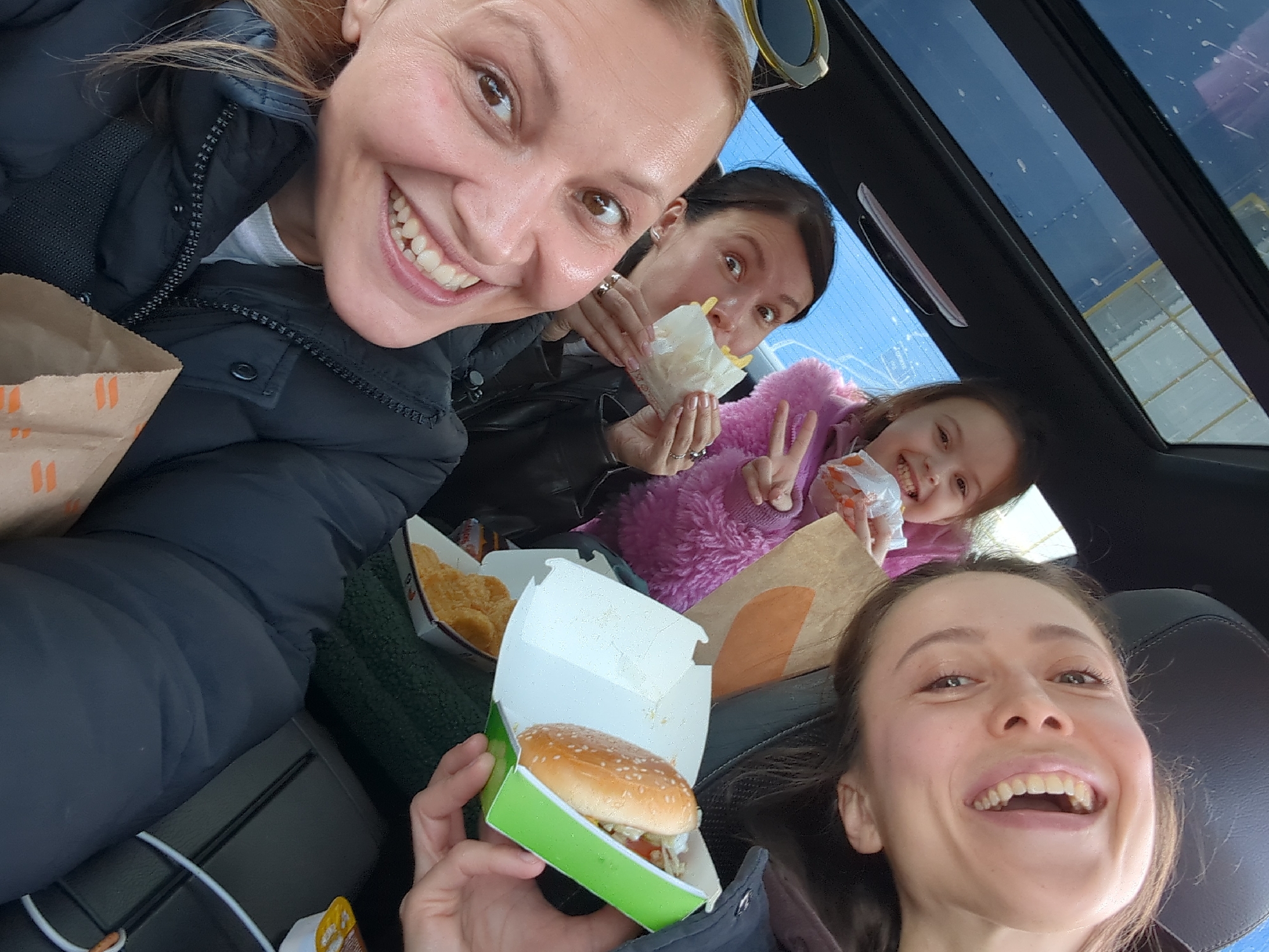 Inside a vehicle in the city of Khimki, Russia, four females are depicted consuming fast food. An adult female with blonde hair occupies the foreground left, wearing a black puffer jacket and a light-colored shirt, smiling widely towards the camera. She holds a brown paper bag with orange stripe branding. To her left, another adult female with dark hair, wearing a black jacket, holds a white paper wrapper containing French fries near her mouth, smiling with wide eyes. Multiple rings are visible on her fingers. A young female child in a bright pink fuzzy jacket is positioned to the right of the second adult, smiling and holding a white paper wrapper with food, while extending her right hand in a peace sign gesture. The fourth individual, an adult female with dark hair, is visible on the far right, head tilted, smiling broadly while holding an open green and white cardboard box containing a hamburger. Below the hamburger box, a white box with chicken nuggets is partially visible. The vehicle's interior features dark upholstery, and a sunroof above reveals a clear blue daytime sky. A pair of sunglasses with a gold-colored frame rests near the top left. All subjects appear cheerful and engaged in eating.