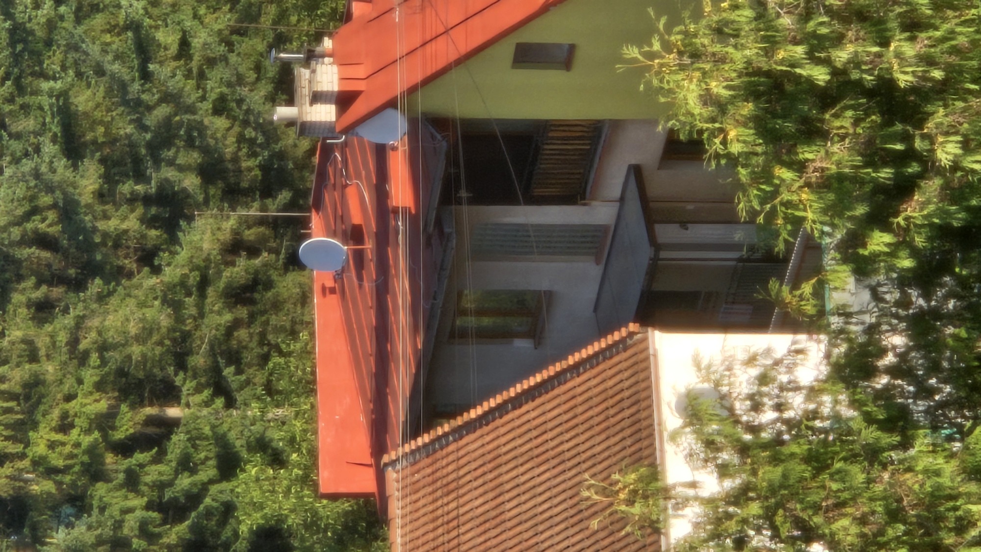 This is a photograph of residential buildings with red tile roofs, set against a backdrop of dense green trees. The image captures an elevated view, suggesting it was taken from a higher vantage point overlooking the neighborhood. In the foreground, lush green trees with feathery foliage partially obscure the view of the houses. The buildings themselves appear to be two to three stories high, with varied architectural styles.
One building features a prominent red roof with visible gutters and several antennas. Adjacent to it, another structure displays a light beige facade with several windows and a balcony. Further to the right, a building with a bright yellow-green side and a red roof is visible. The roofs are punctuated by chimneys, and satellite dishes are mounted on some of the red roofs. The overall impression is of a residential area nestled within a wooded environment. The lighting suggests it is either early morning or late afternoon, with sunlight casting shadows and highlighting the textures of the trees and rooftops.
There are no people visible in the image, and no specific actions or interactions are depicted. The scene is static and focused on the architectural and natural elements of the environment. The presence of multiple buildings and their proximity to the dense tree line indicate a suburban or semi-rural setting. Given the visual cues, this could be a residential area in Prešov, Slovakia, known for its proximity to natural landscapes and green spaces.