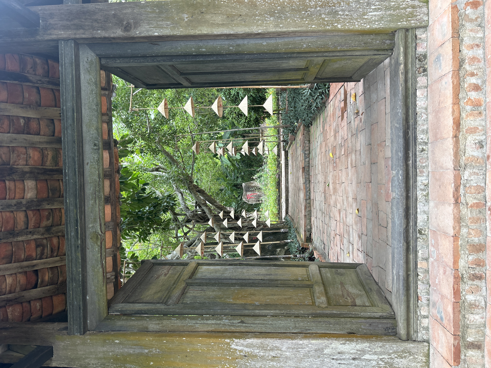 This is a photograph of a pathway leading through an open wooden doorway. The doorway is made of aged, weathered wood and is flanked by two large wooden doors, one of which is slightly ajar. Above the doorway is a tiled roof made of weathered, terracotta-colored tiles. The pathway is paved with red bricks and is lined with green foliage. Suspended from branches of a large tree are numerous conical straw hats, appearing to be decorative items. In the background, a large rock with red markings is visible, and the area is densely covered with green vegetation. The scene suggests an outdoor location, possibly a garden or a temple, within a tropical environment. The overall impression is one of rustic charm and natural beauty. The style of the architecture and the presence of the conical hats are indicative of Vietnamese culture. This scene likely depicts a part of the Long Son Pagoda in Nha Trang, Vietnam, given its architectural style and the presence of similar decorative elements.