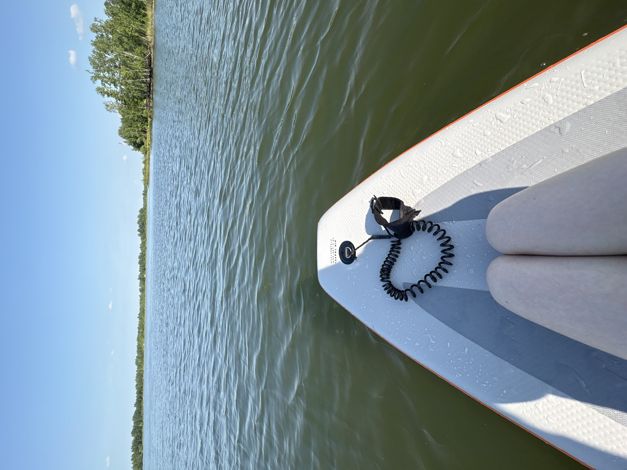 The image shows a point-of-view shot from a paddleboard on a body of water. In the foreground, the front of the paddleboard is visible, with a coiled leash and a person's legs. The water is calm with small ripples, and the background features a tree line along the horizon under a clear blue sky. The scene appears to be a natural setting, possibly a lake or a calm river, located in or near the city of Razdolnoye, Russia, given the context of the prompt.