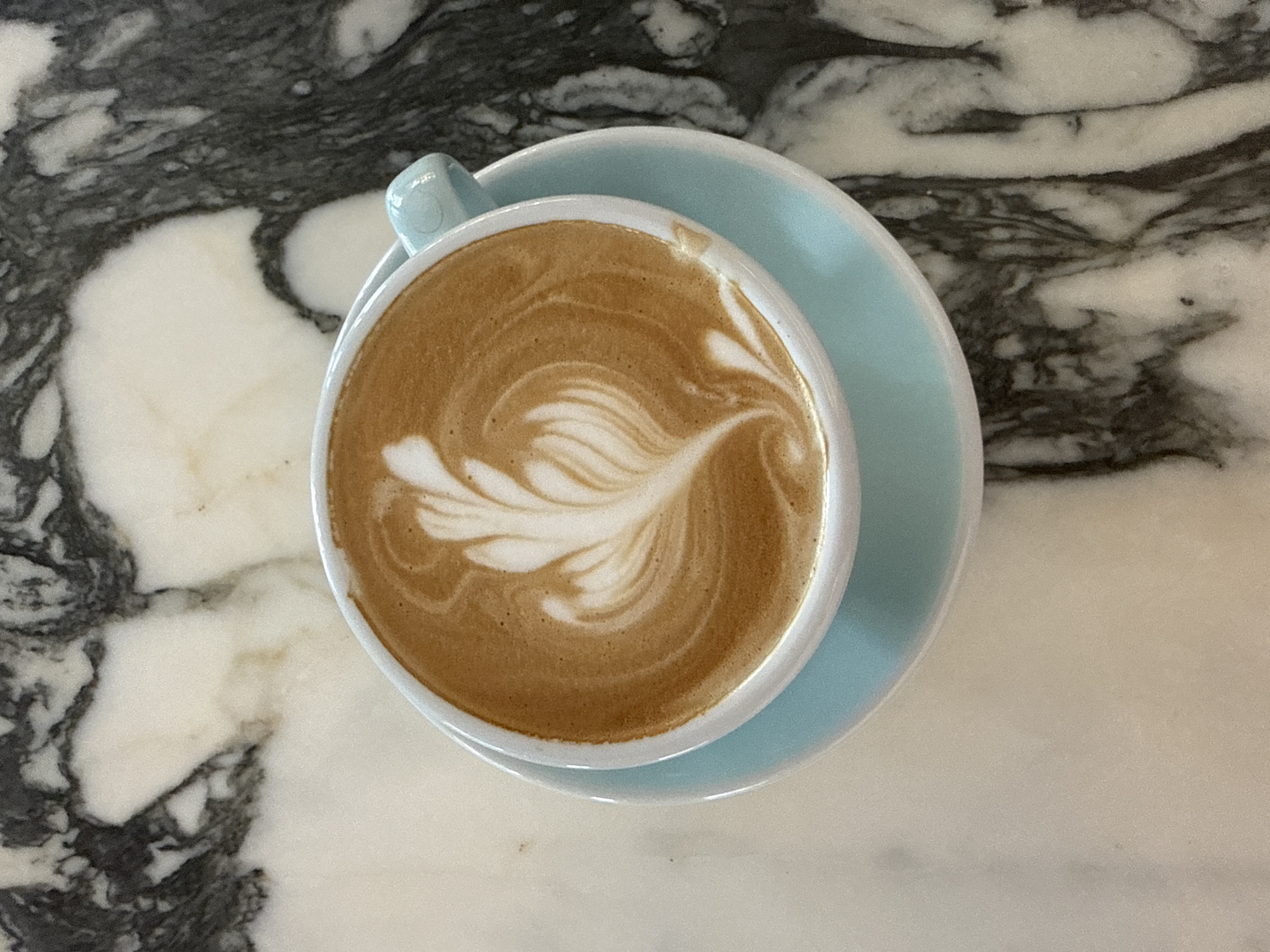 A close-up, overhead shot shows a light blue saucer with a white cup on top, filled with latte art. The cup contains a tan-colored liquid with intricate white foam art resembling a feather or leaf. The cup and saucer are placed on a marble surface with a black and white swirled pattern. The image appears to be taken at a cafe in Hua Hin City Municipality, Thailand.