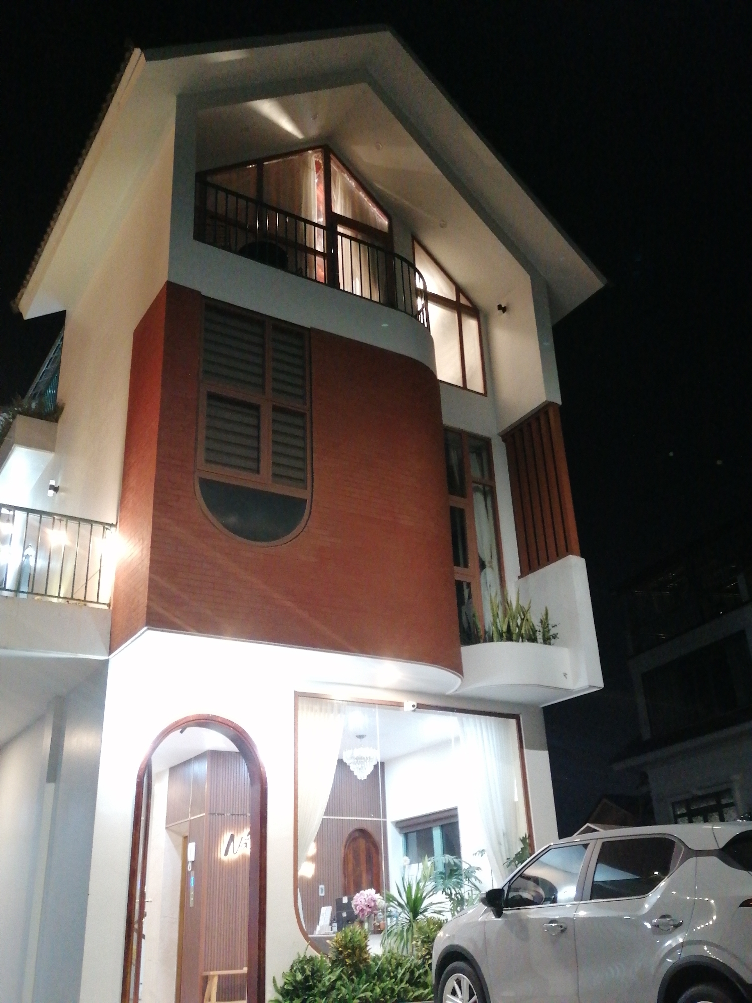 A modern, multi-story building with a striking brick facade and white accents is illuminated against a dark night sky. The architecture features large windows, balconies with railings, and a prominent arched entrance. A light-colored car is parked in front of the building. The scene appears to be on a street in Da Lat, Vietnam, as indicated by signage within the arched doorway that displays Vietnamese text. The overall impression is one of contemporary design and urban nighttime ambiance.